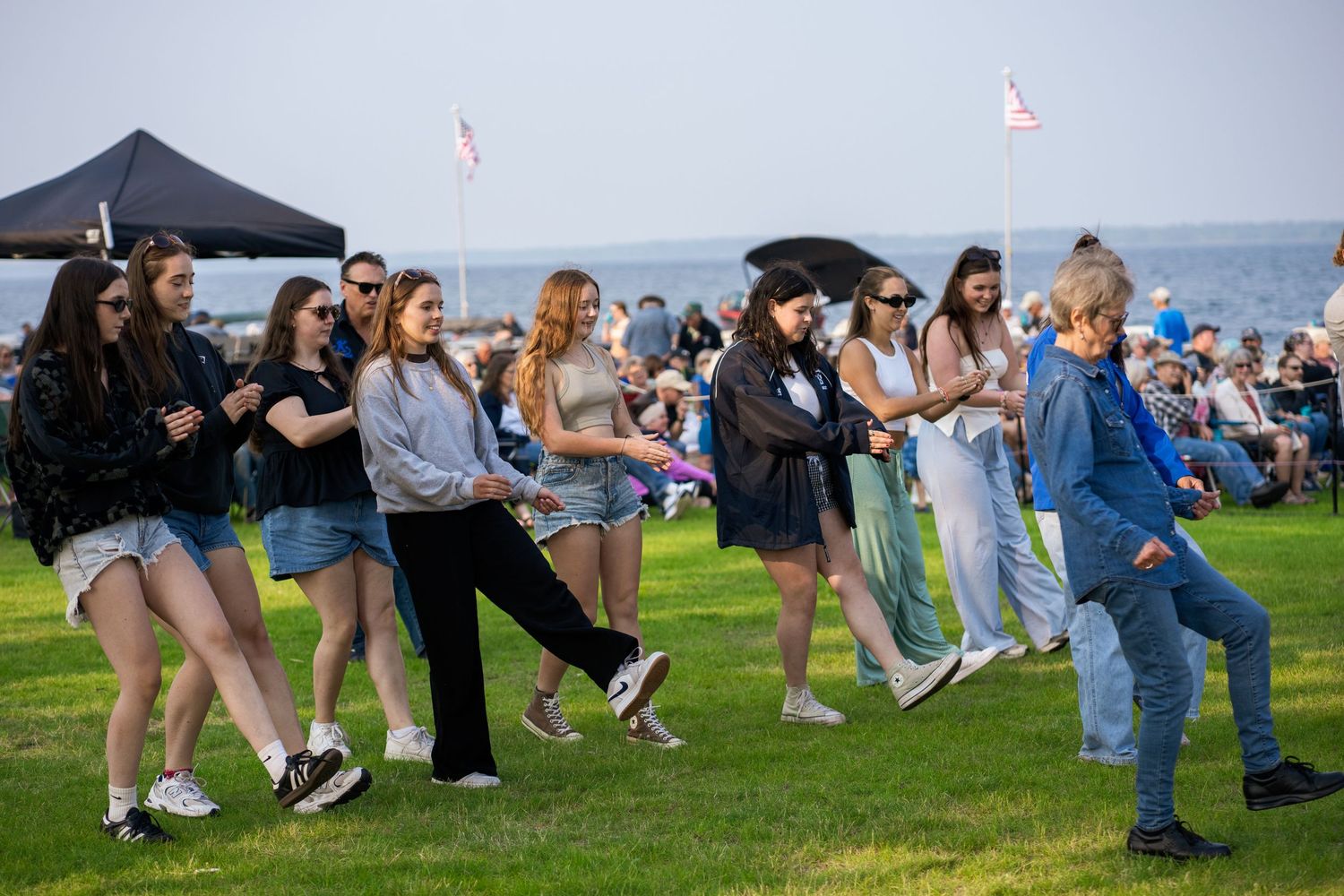 Large group of people dancing on a grassy field near the lake on a sunny day.