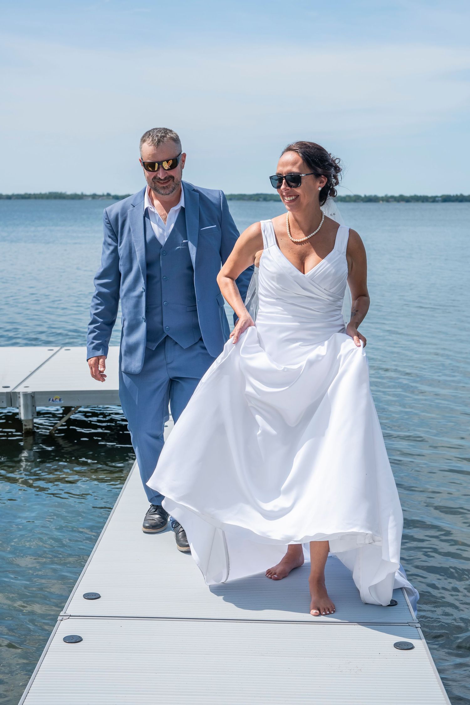 Bride lifts dress while dancing with groom on sunny dock overlooking calm waters.