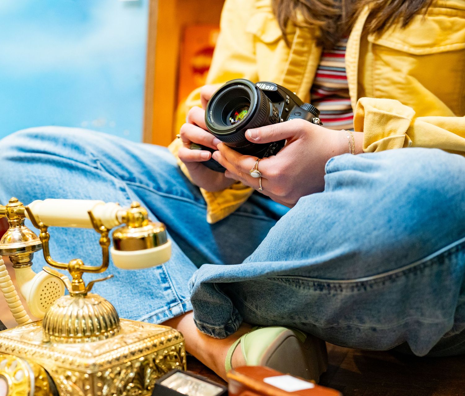 A woman in jeans and yellow jacket holds a DSLR camera beside a ornate gold vintage telephone.