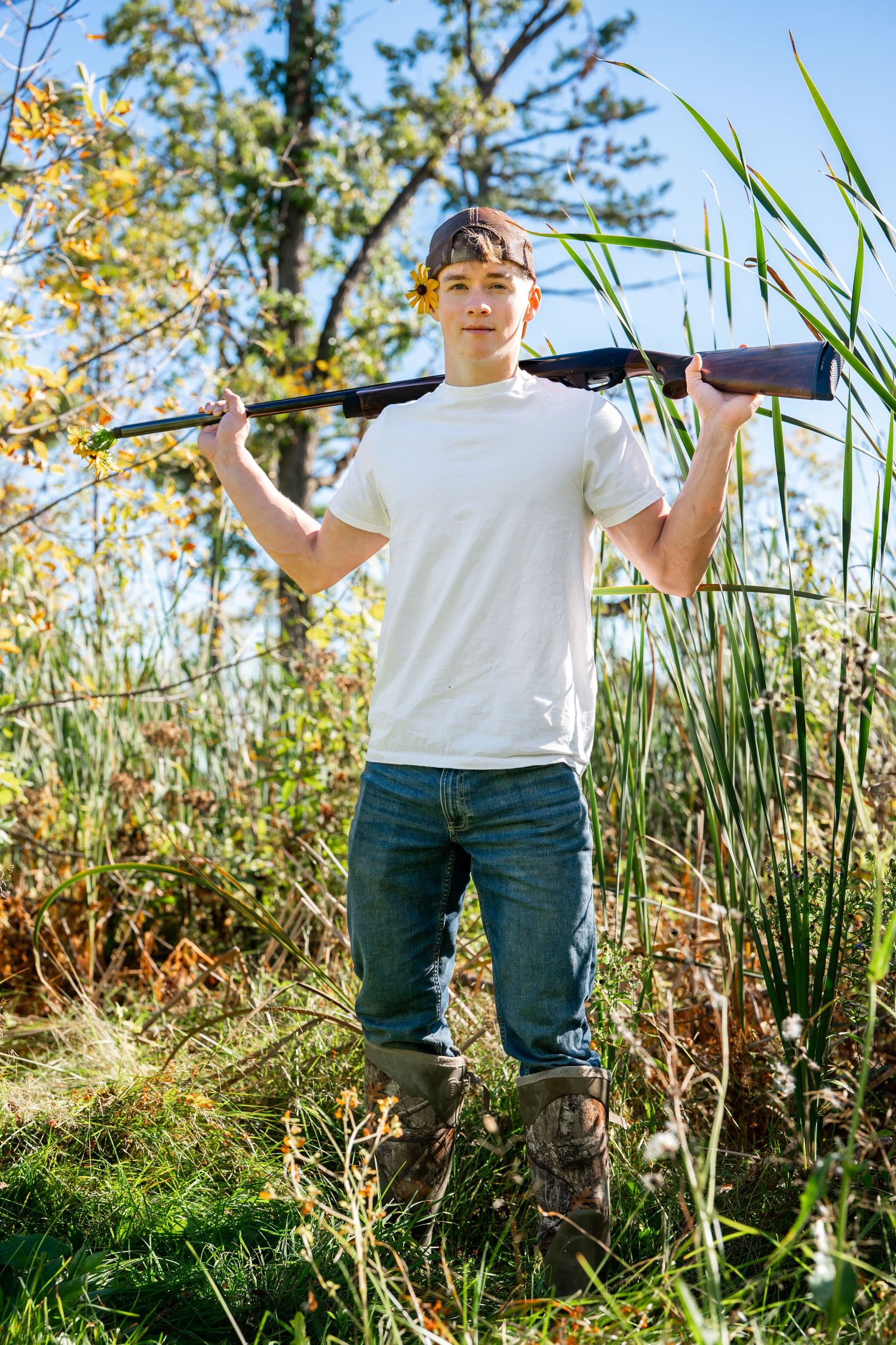 Hunter in white shirt holding rifle across shoulders in tall grass and autumn foliage.