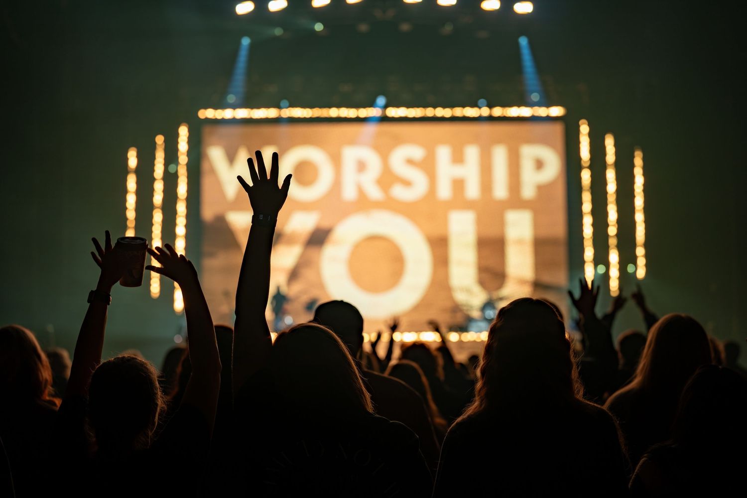 Silhouetted crowd raising hands at worship concert with illuminated 'WORSHIP' text on screen.