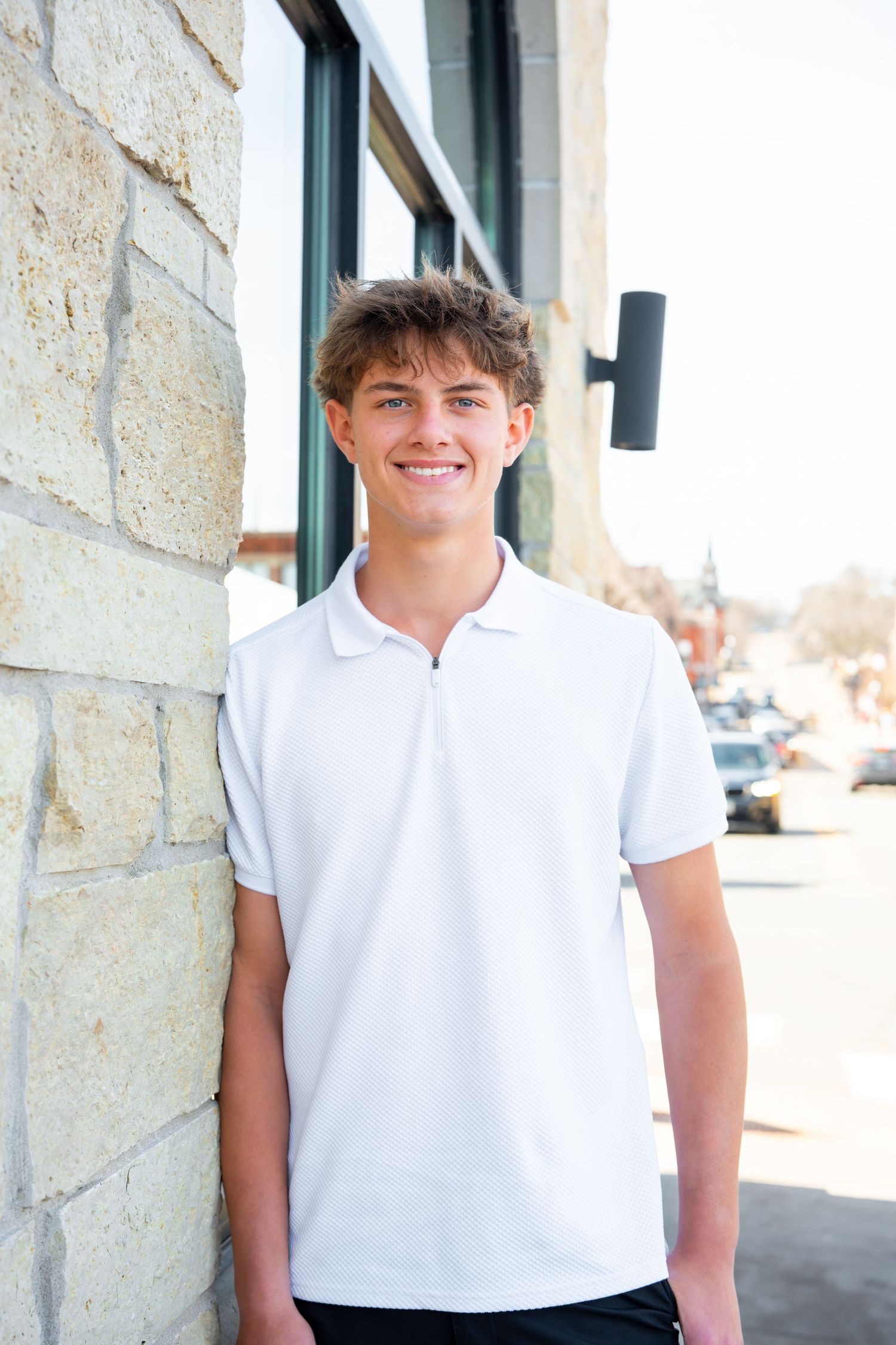 A young person in a white polo shirt stands against a stone wall on a sunny day.