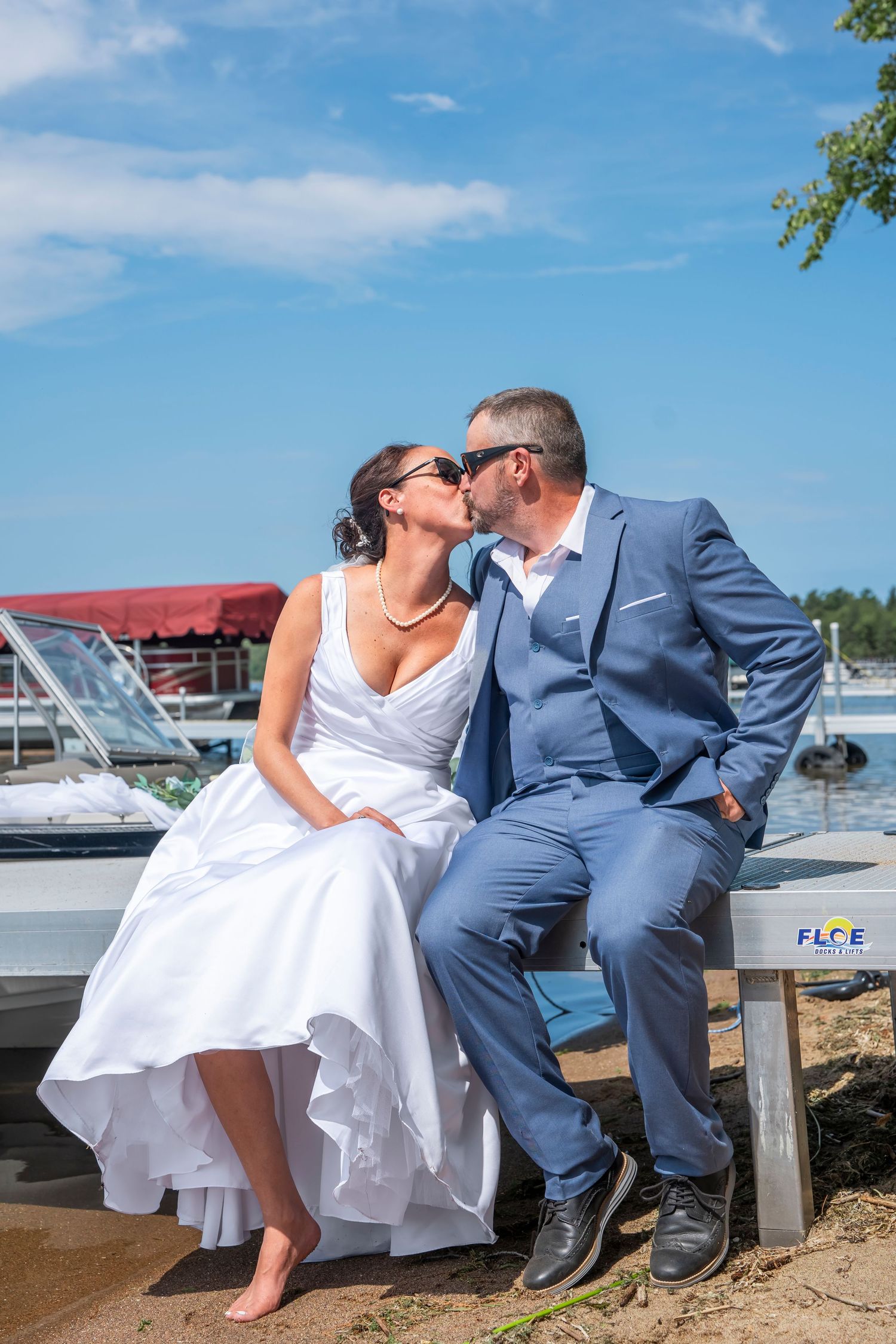 Couple in wedding attire sitting on dock by marina on sunny day.