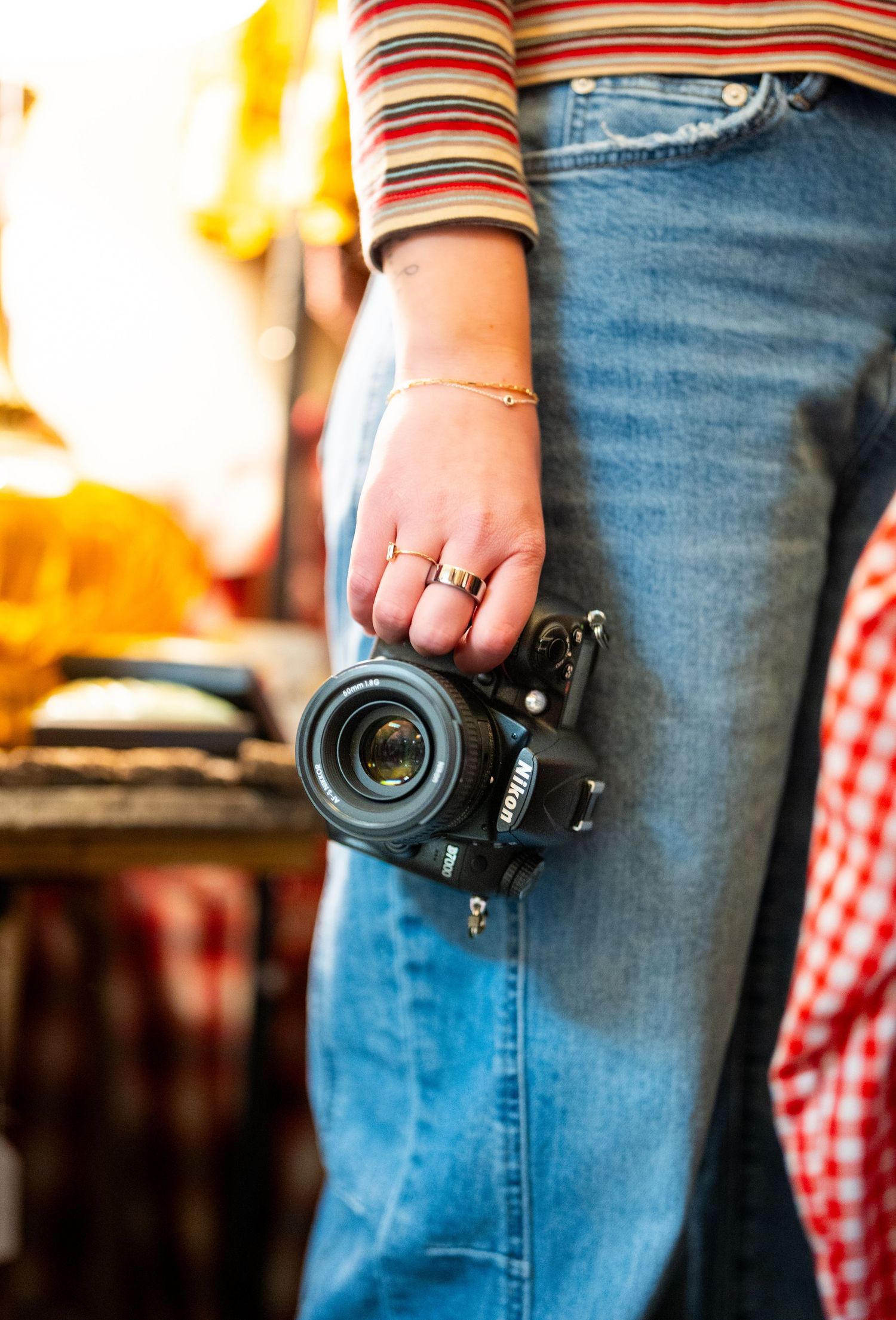 Woman in jeans and striped top holding a Nikon DSLR camera with gold rings and bracelet in a vintage shop.