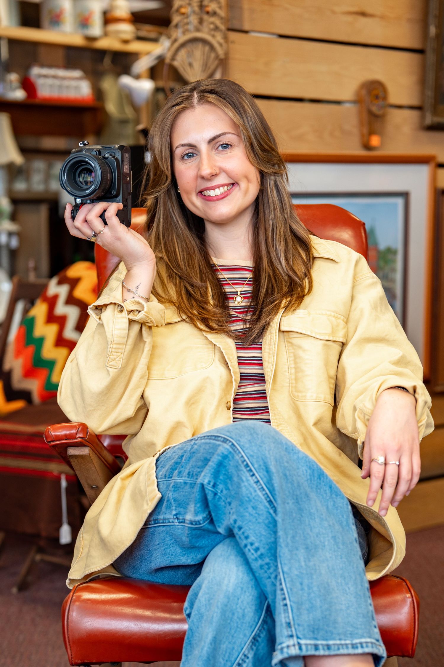 Smiling woman in yellow jacket holding vintage camera seated in red leather chair surrounded by retro decor.