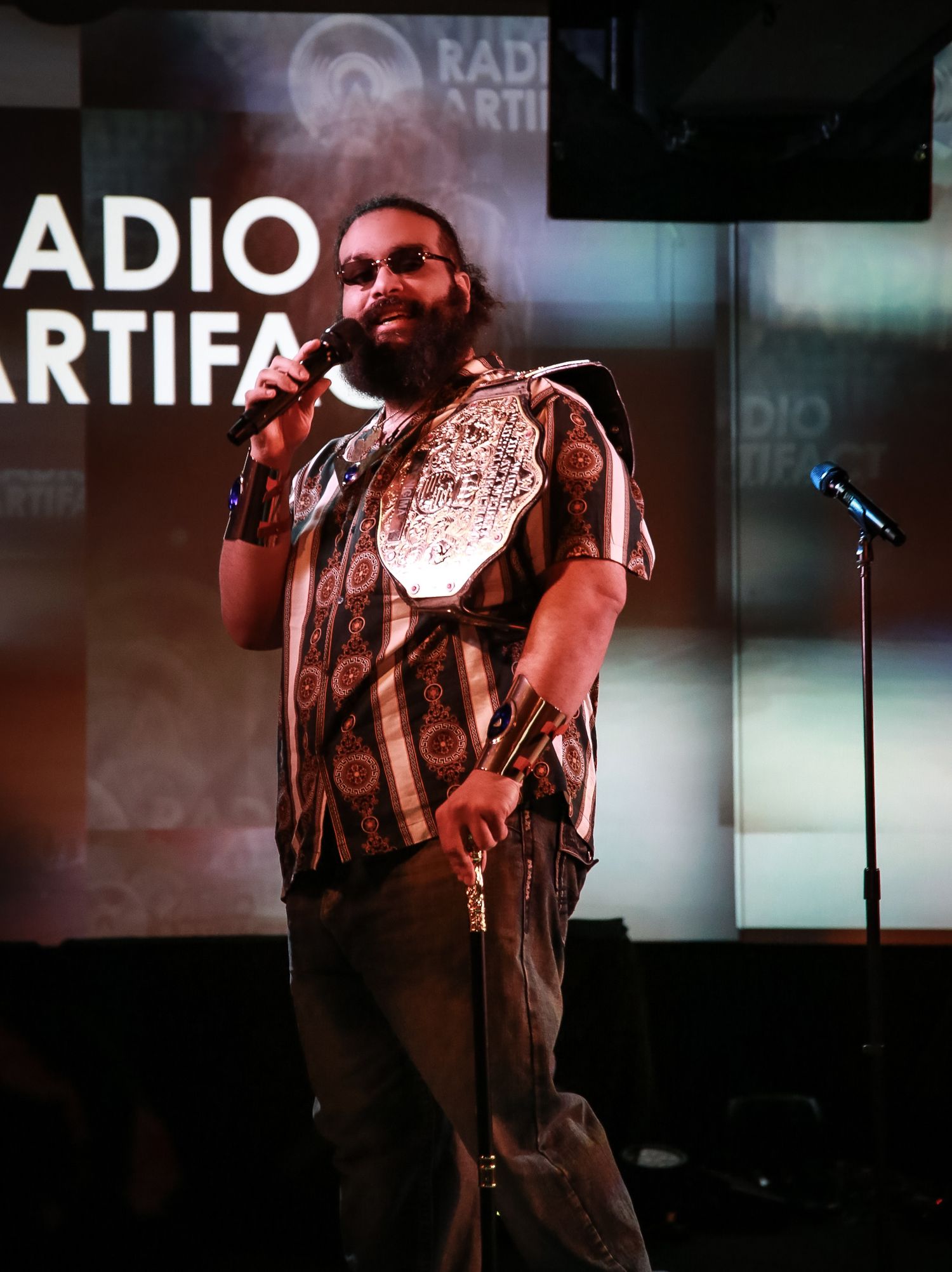 Performer wearing sunglasses and patterned shirt stands on stage at Radio ARTIFA with microphone stand visible.