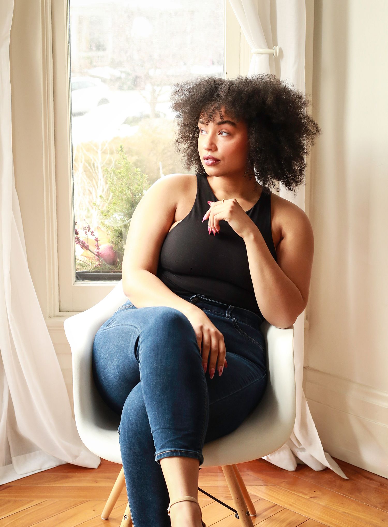 A person in a black tank top and jeans sits thoughtfully in a white modern chair by a sunlit window with sheer curtains.