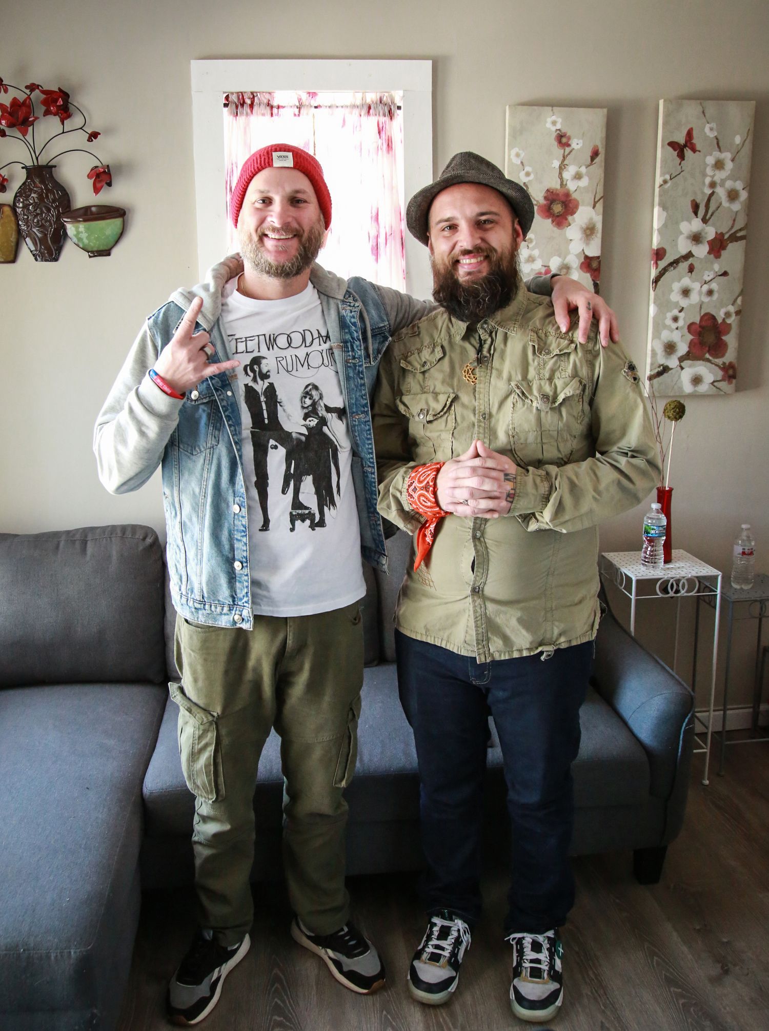 Two friends wearing casual clothing and beanie hats stand together in a living room interior with wall decorations.