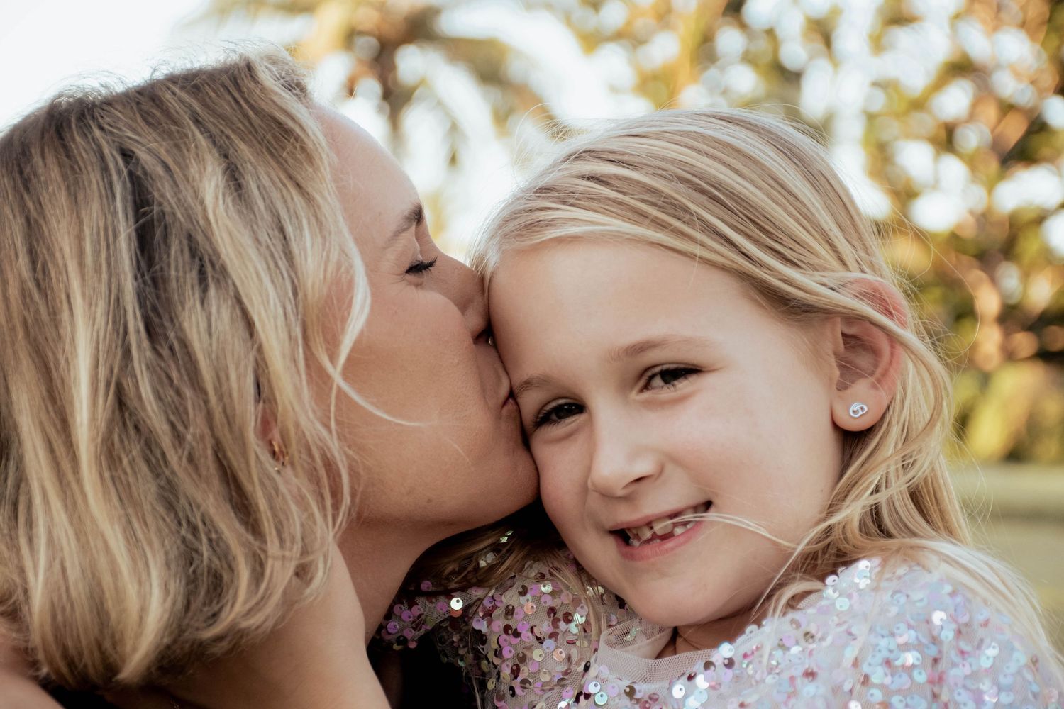 A mother lovingly kissing her smiling young blonde daughter on the cheek outdoors.