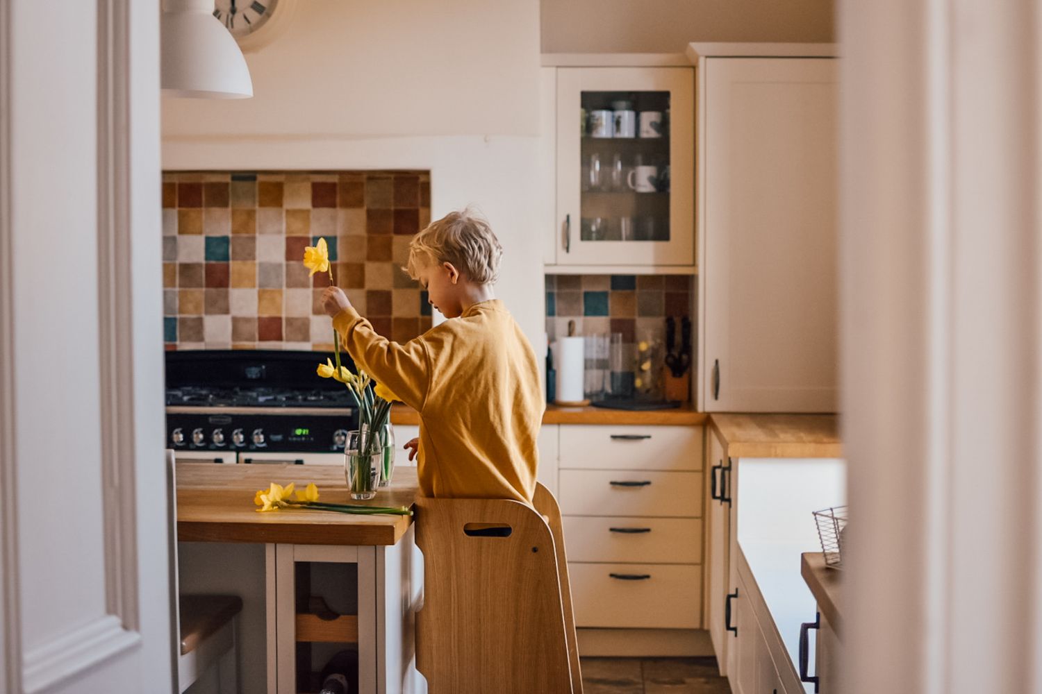 Boy standing on a kitchen helper putting daffodils in a vase.