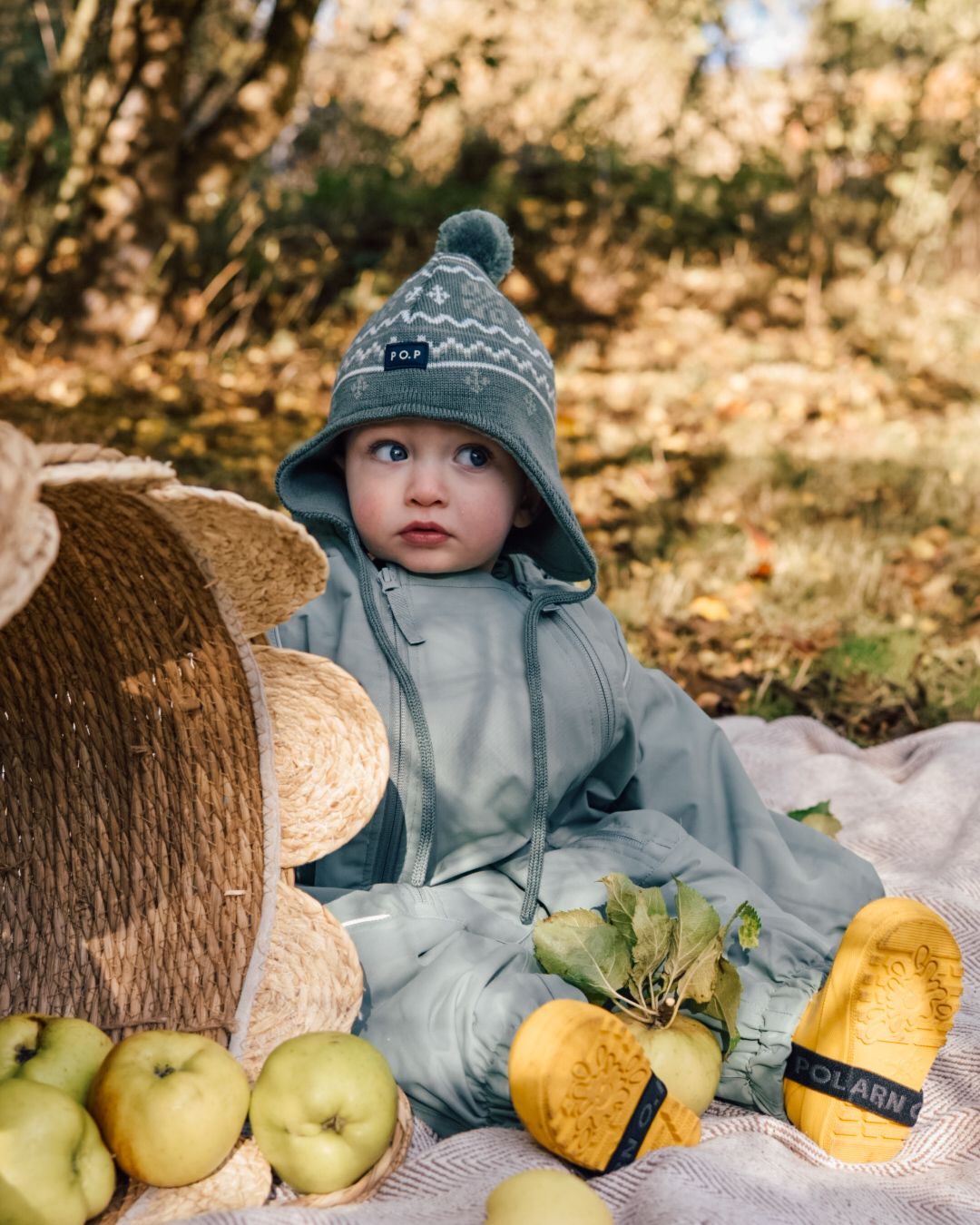 Baby sat on blanket in winter atire next to a tipped over basket spilling green apples out.
