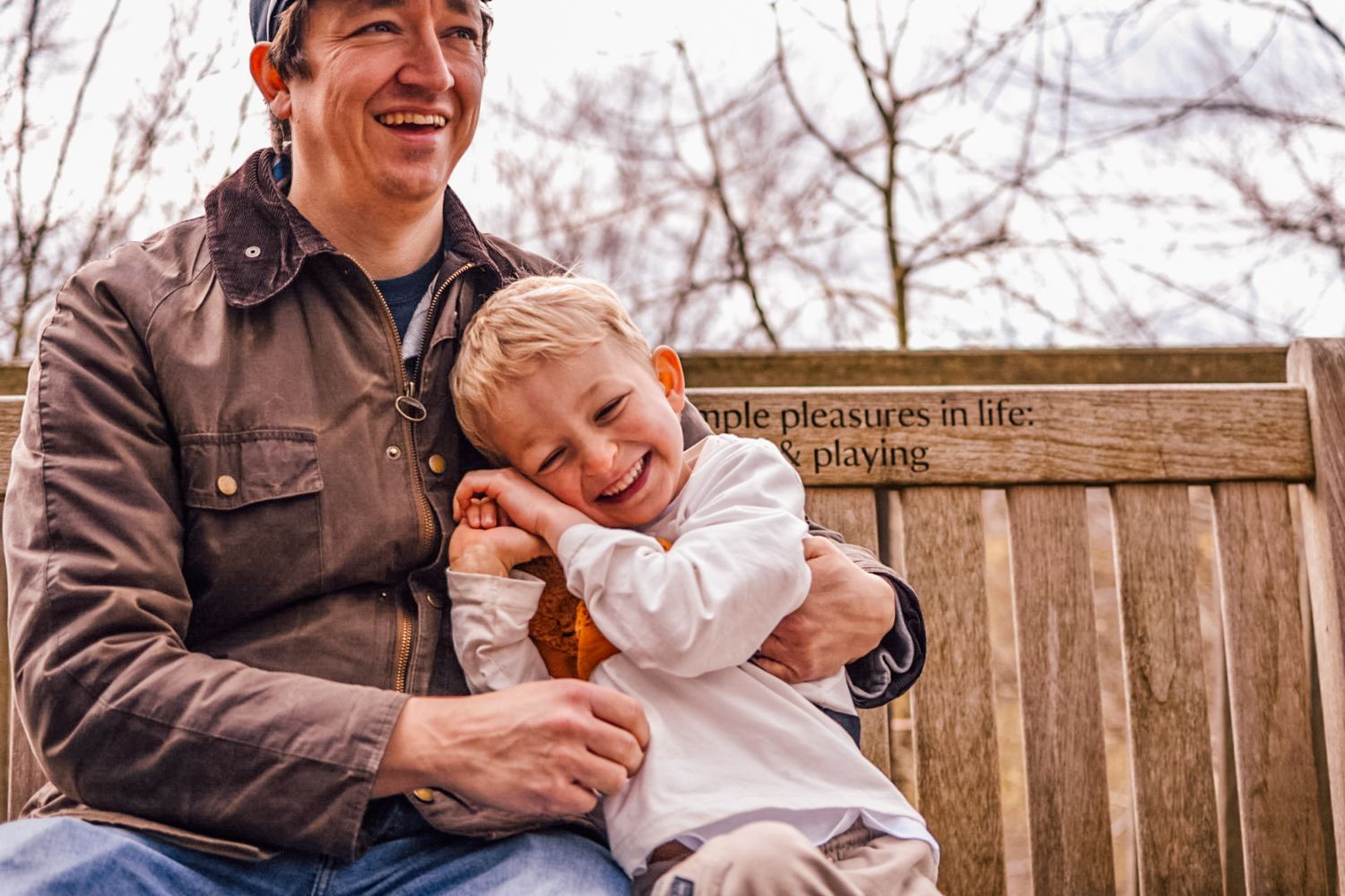 Man and boy sat on wooden outdoor bench both smiling and hugging.