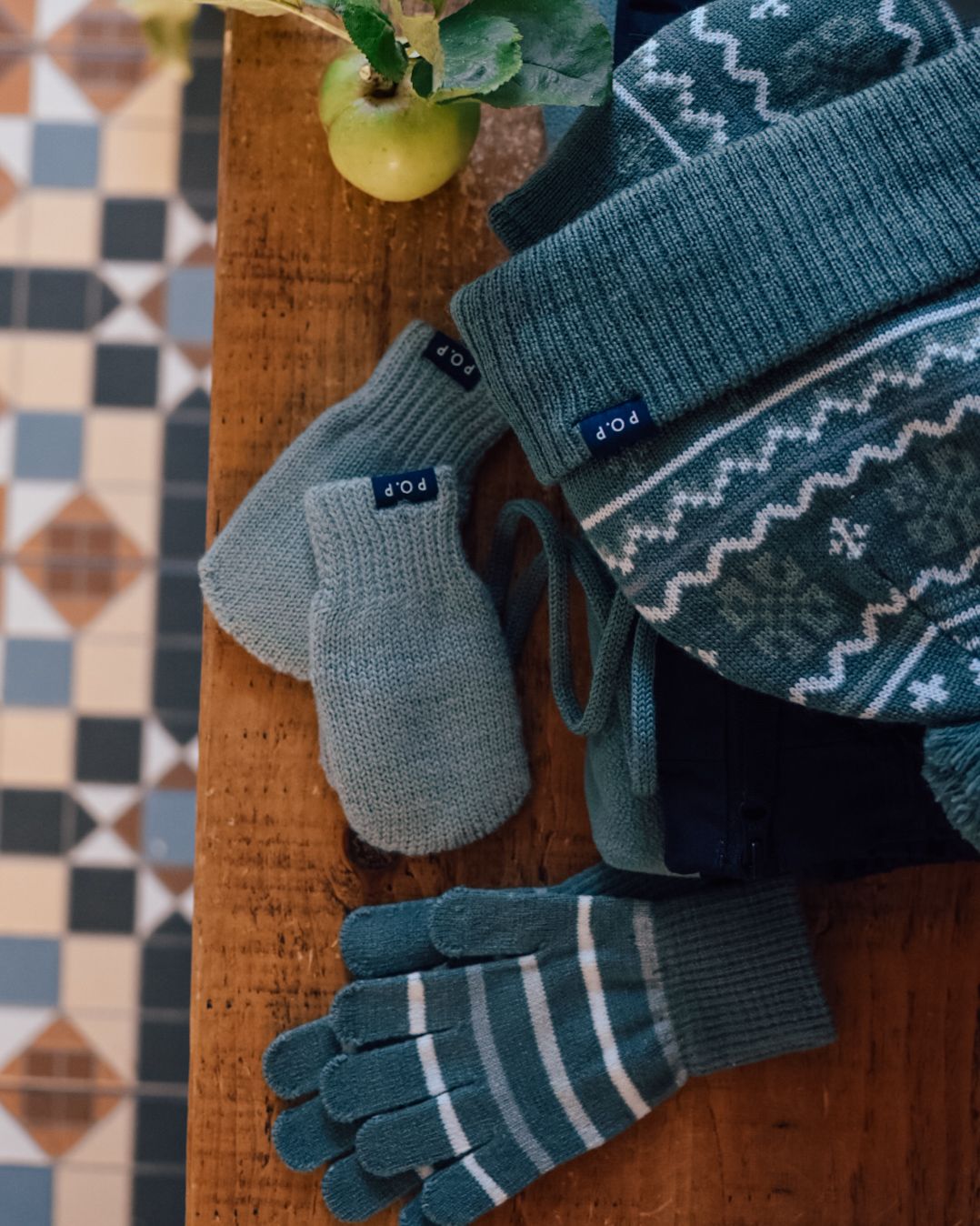 Wooly hat, gloves and mittens on a wooden top with coloured floor tiles in the background.