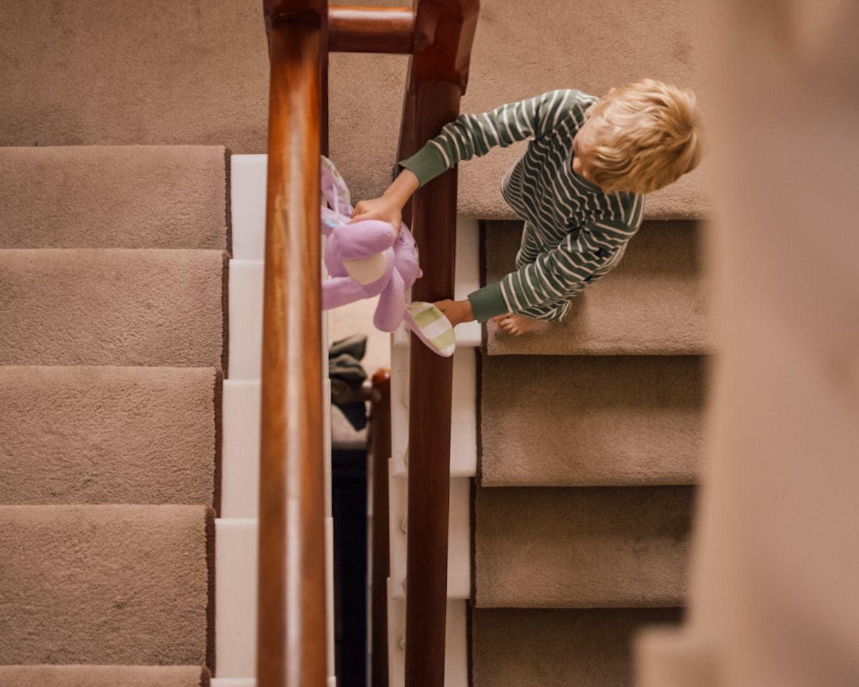 Boy playing with pink soft toy bunny rabbit on large carpeted staircase with wooden hand rail