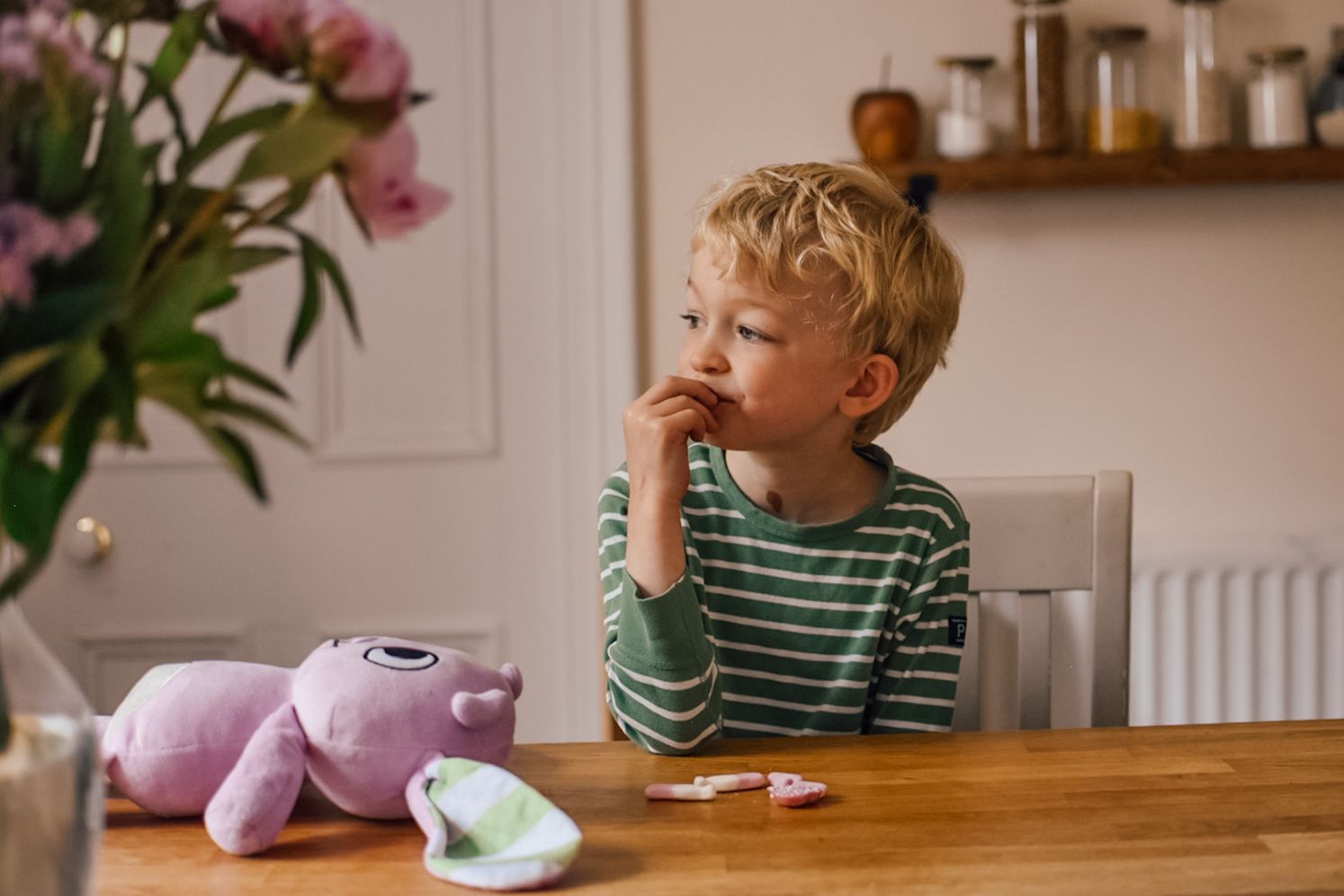Boy eating whilst sat at breakfast bar with pink soft toy bunny in front of him and bokeh image of flowers in foreground