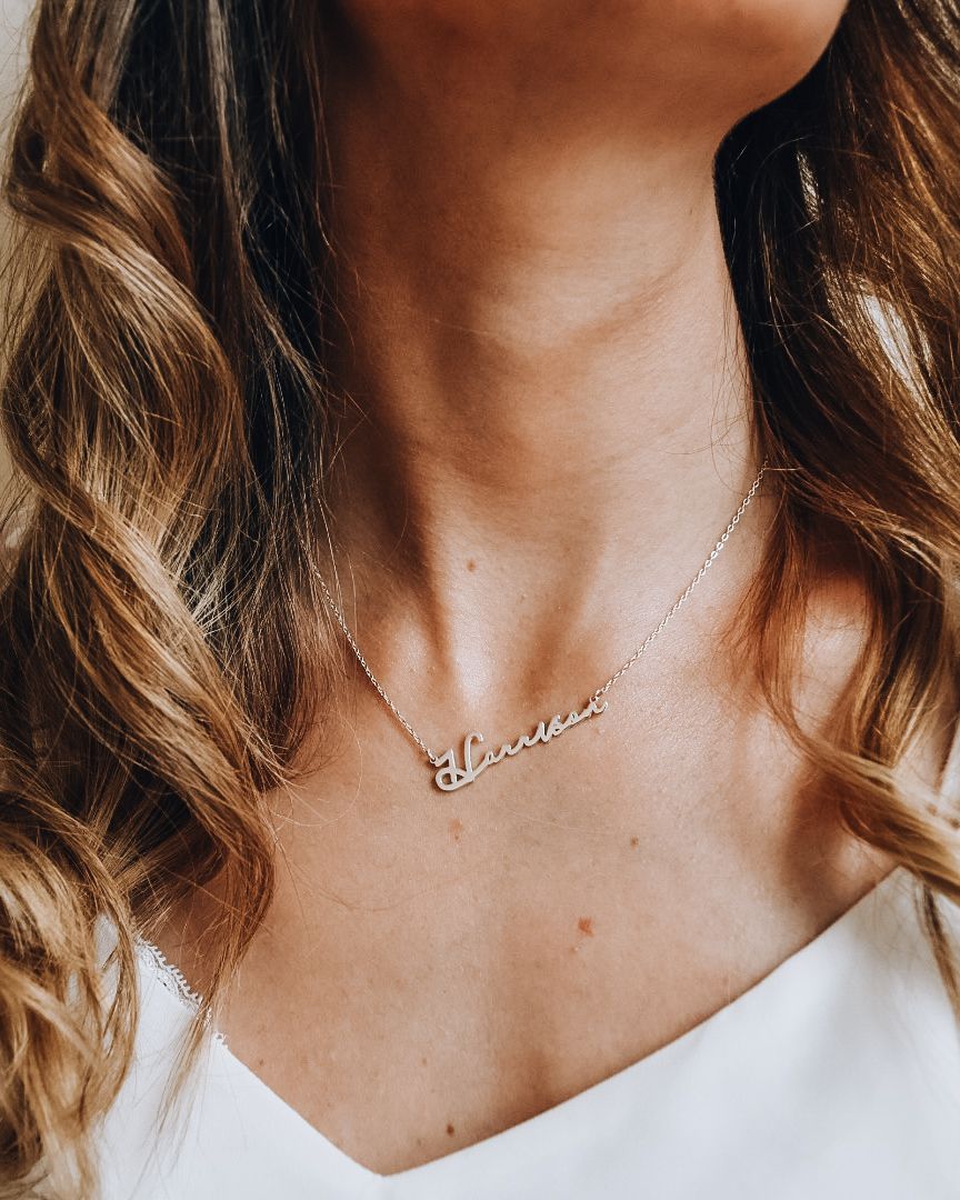 Lady with brown hair wearing a white top and a silver necklace with the name 