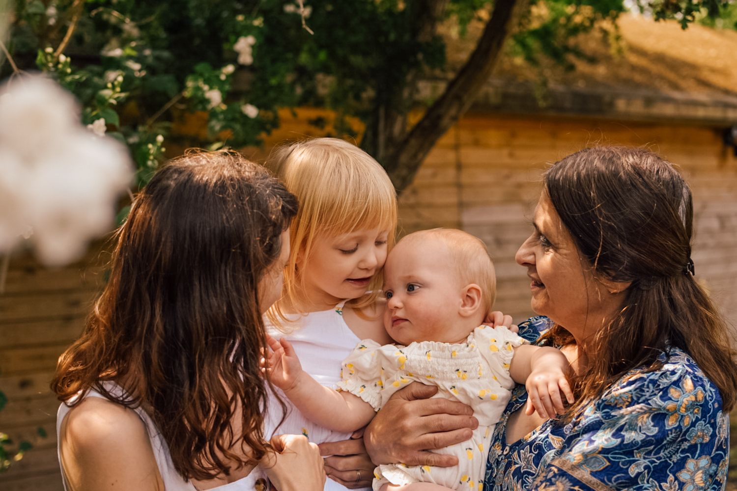 Mother, girl, baby and Grandmother embrace together in front of garden shed and trees