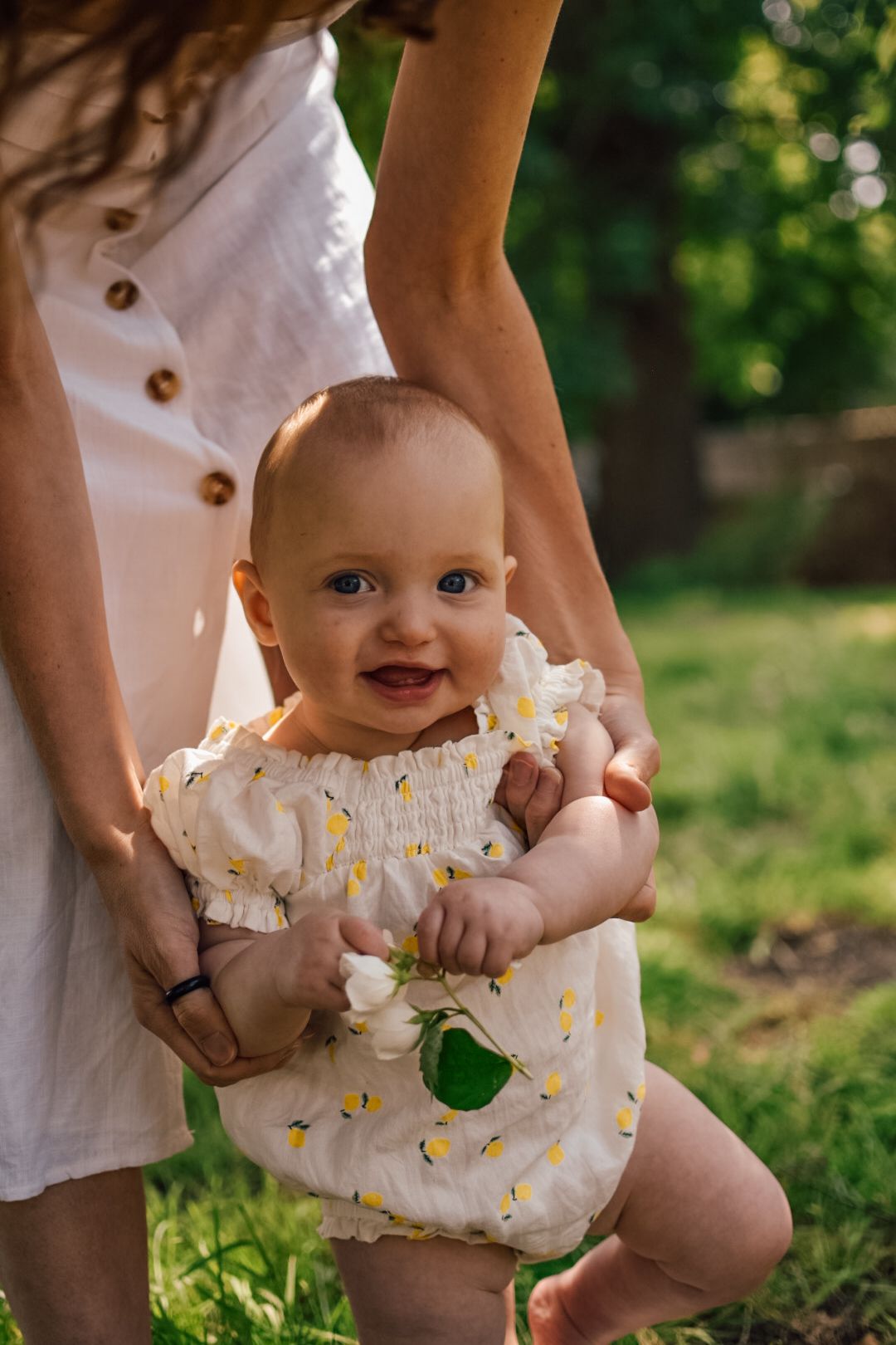 Arms of mother holding baby whilst she walks in the garden holding tree blossom in her hands.