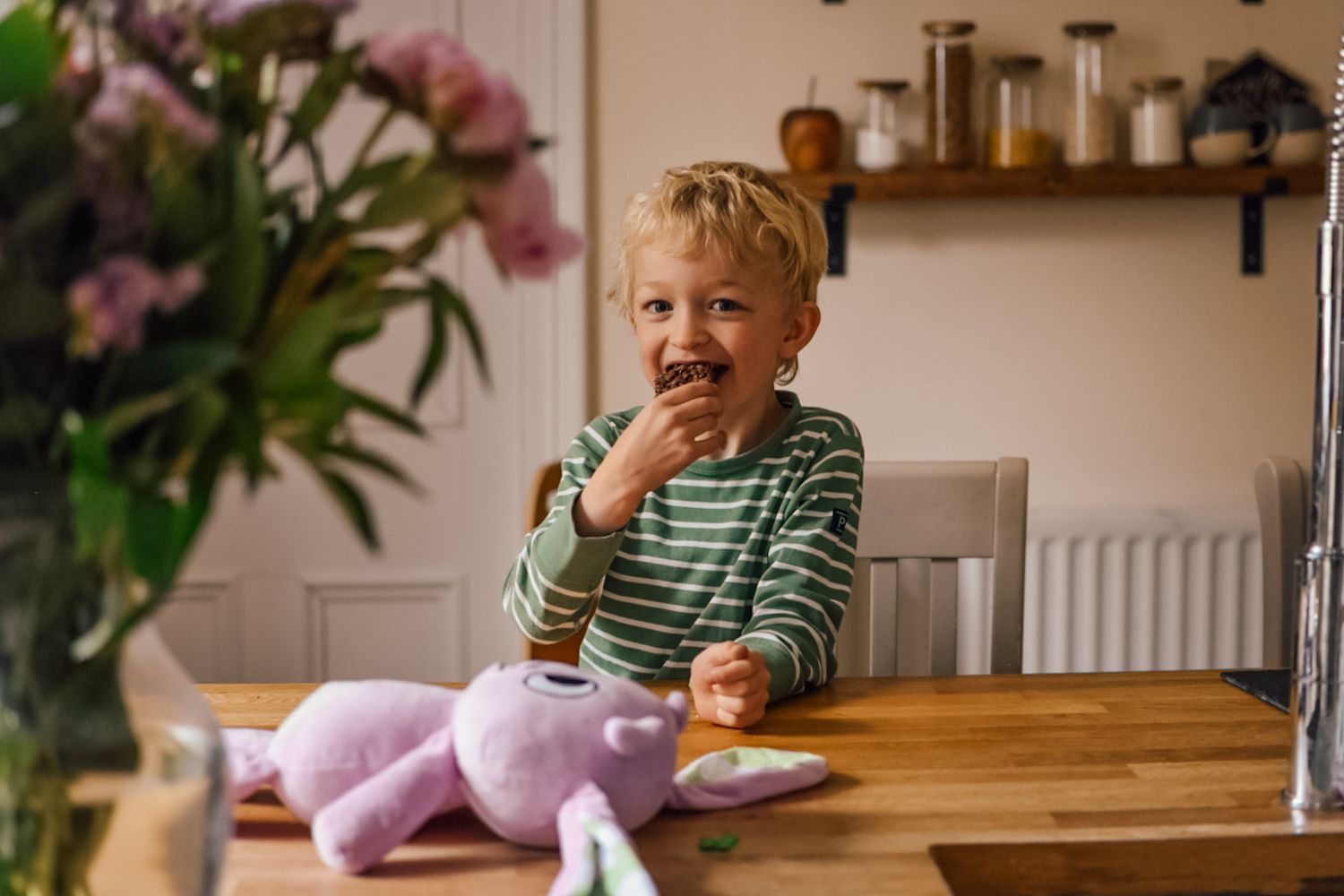 Boy eating whilst sat at breakfast bar with pink soft toy bunny in front of him and bokeh image of flowers in foreground