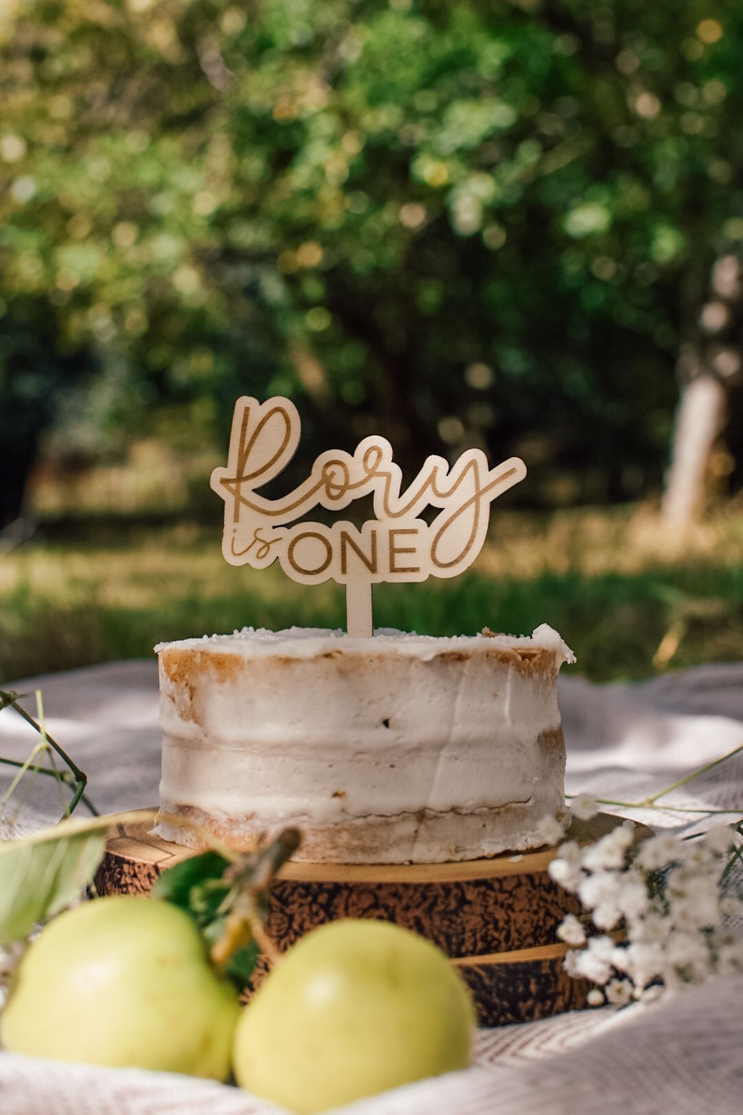 Cake with a wooden topper reading 'Rory, One' with green apples in the foreground, on top of a blanket with wooded area in the background.