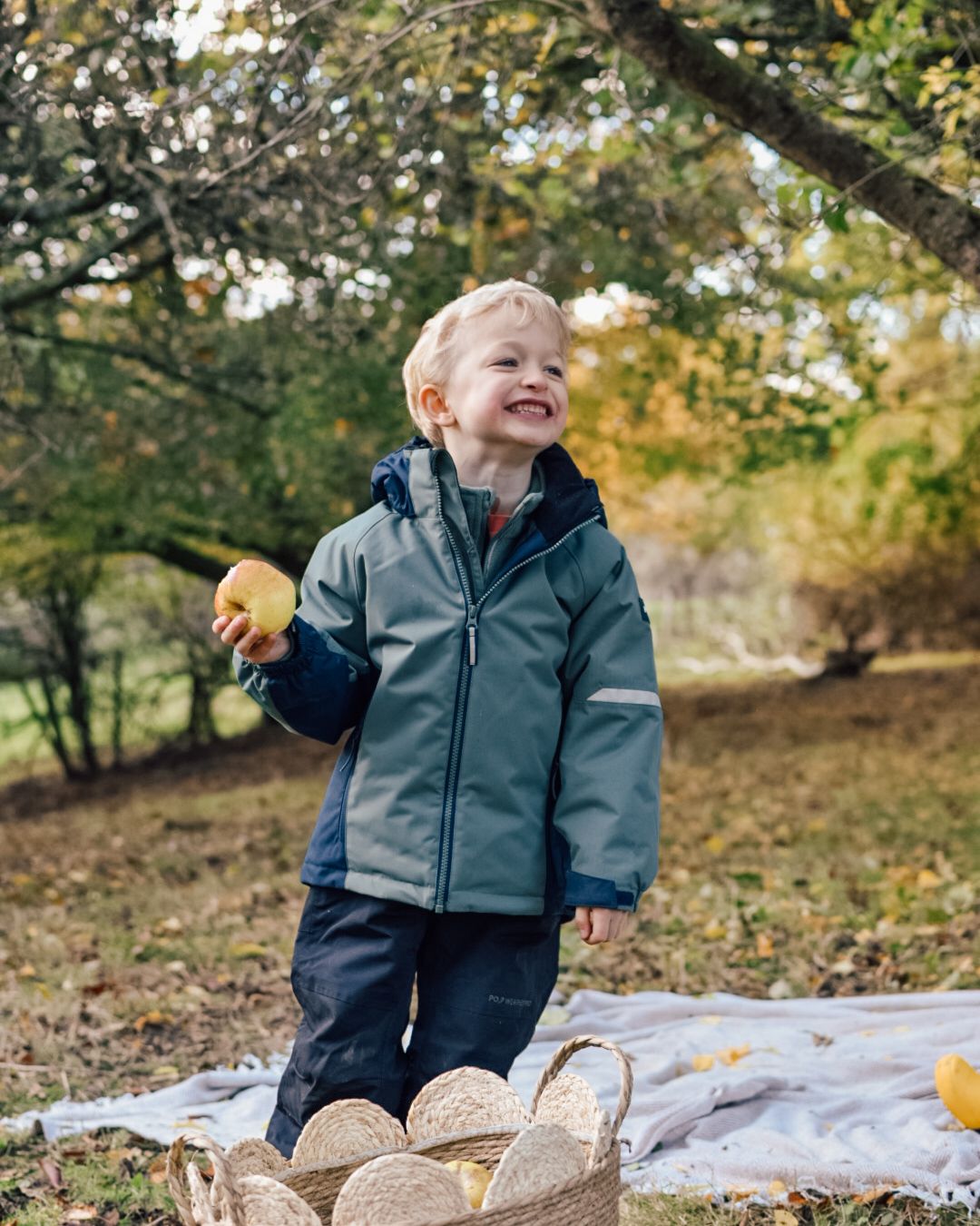 Boy in warm outdoor clothing holding an apple picked from trees in woodland scene.