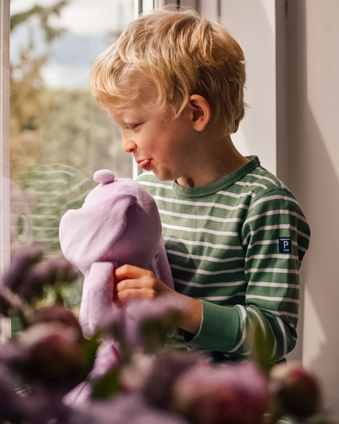 Boy holding pink soft toy on window ledge looking out with bokeh effect flowers in foreground