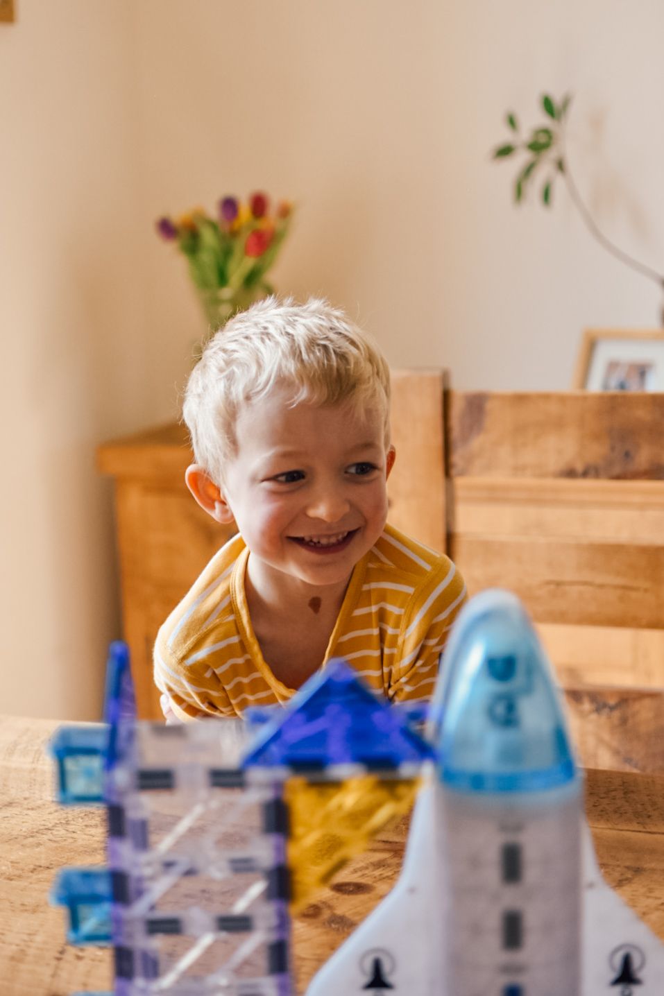 Boy playing with magna tiles space ship product on a wooden table sat at a wooden chair with a wooden sideboard in the background