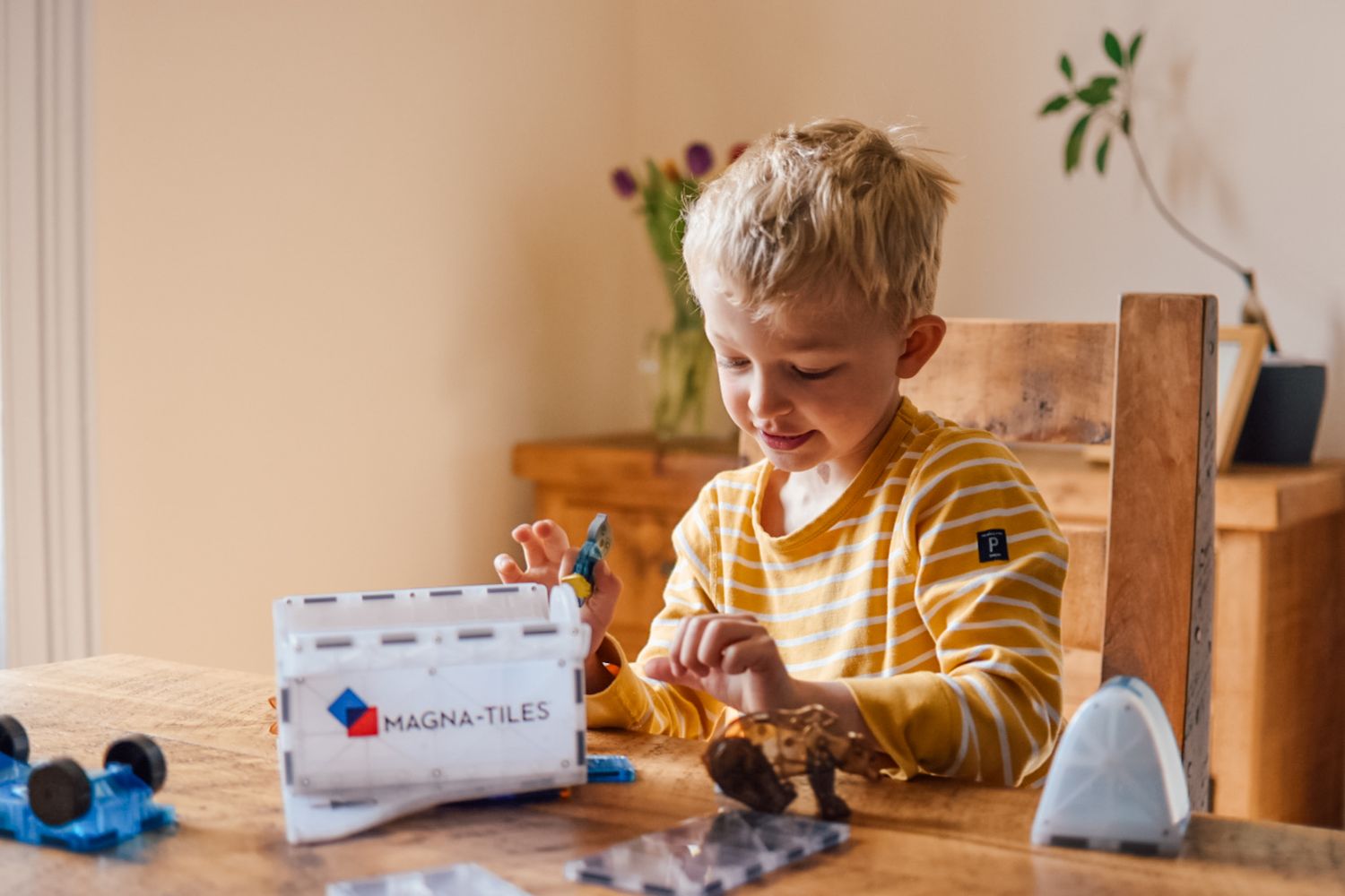 Boy playing with magna tiles space ship product on a wooden table sat at a wooden chair with a wooden sideboard in the background