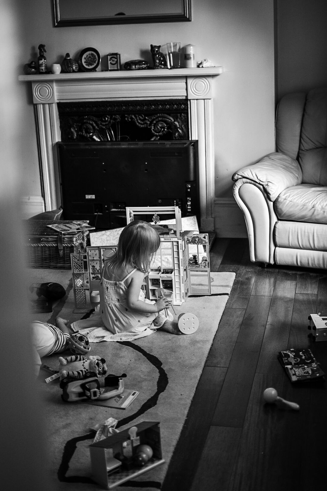 Black and white photo of girl playing with toys on a rug on a wooden floor with fireplace and chair in background.
