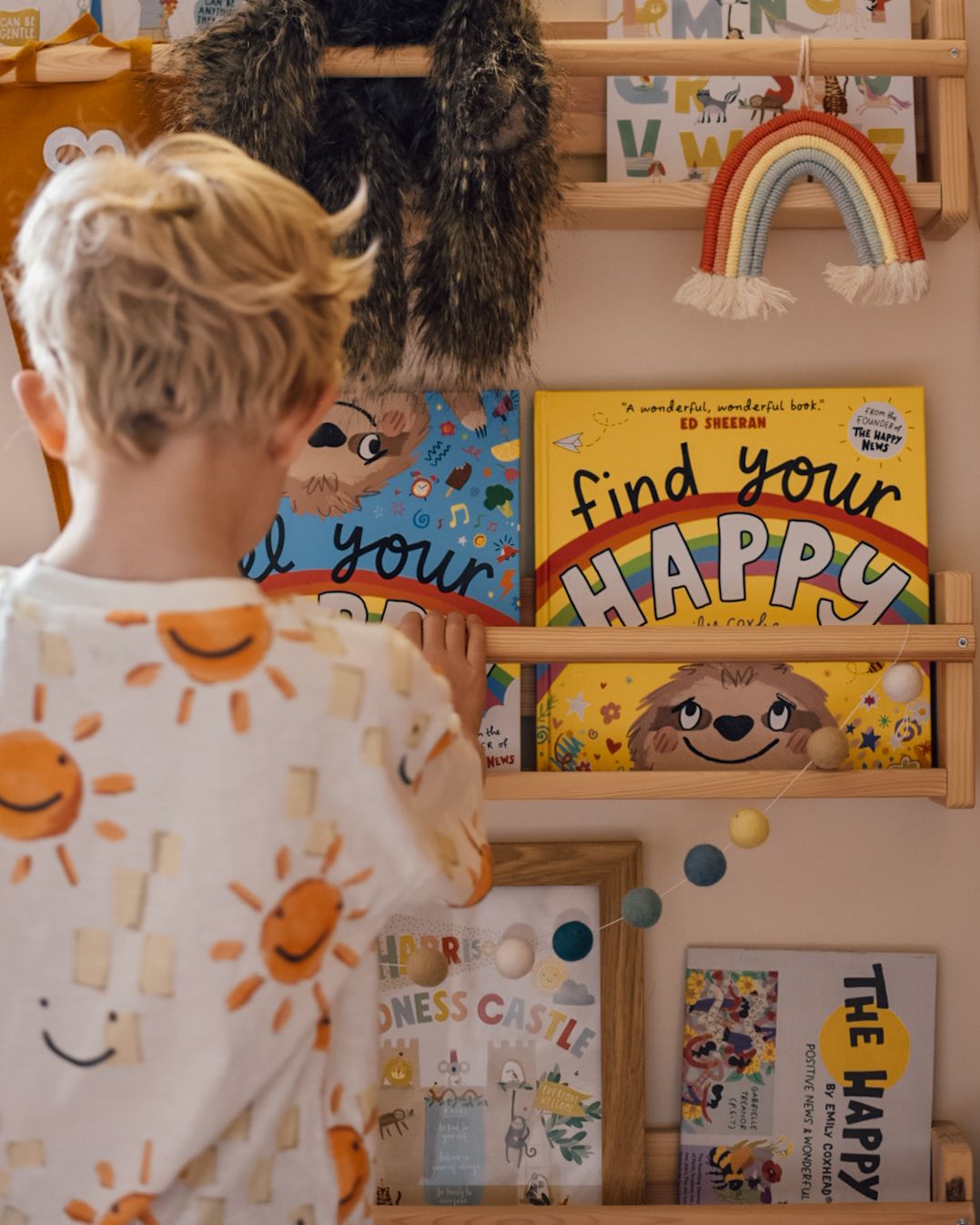 Boy looking at bookcase full of colourful rainbow books. With find your happy book by Emily Coxhead visible