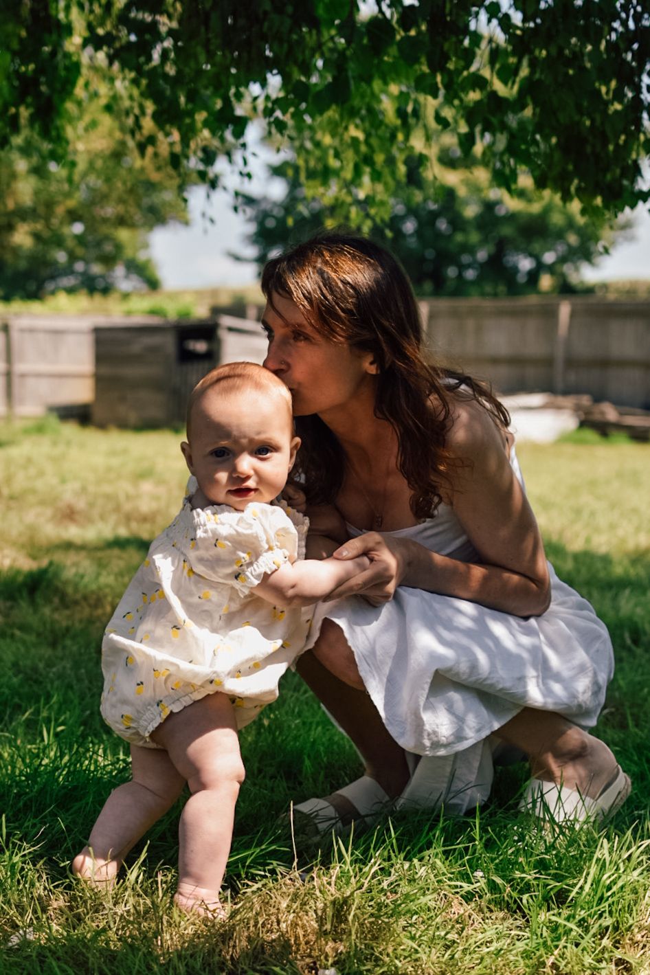 Mother and baby embrace whilst kneeling and standing in garden scene