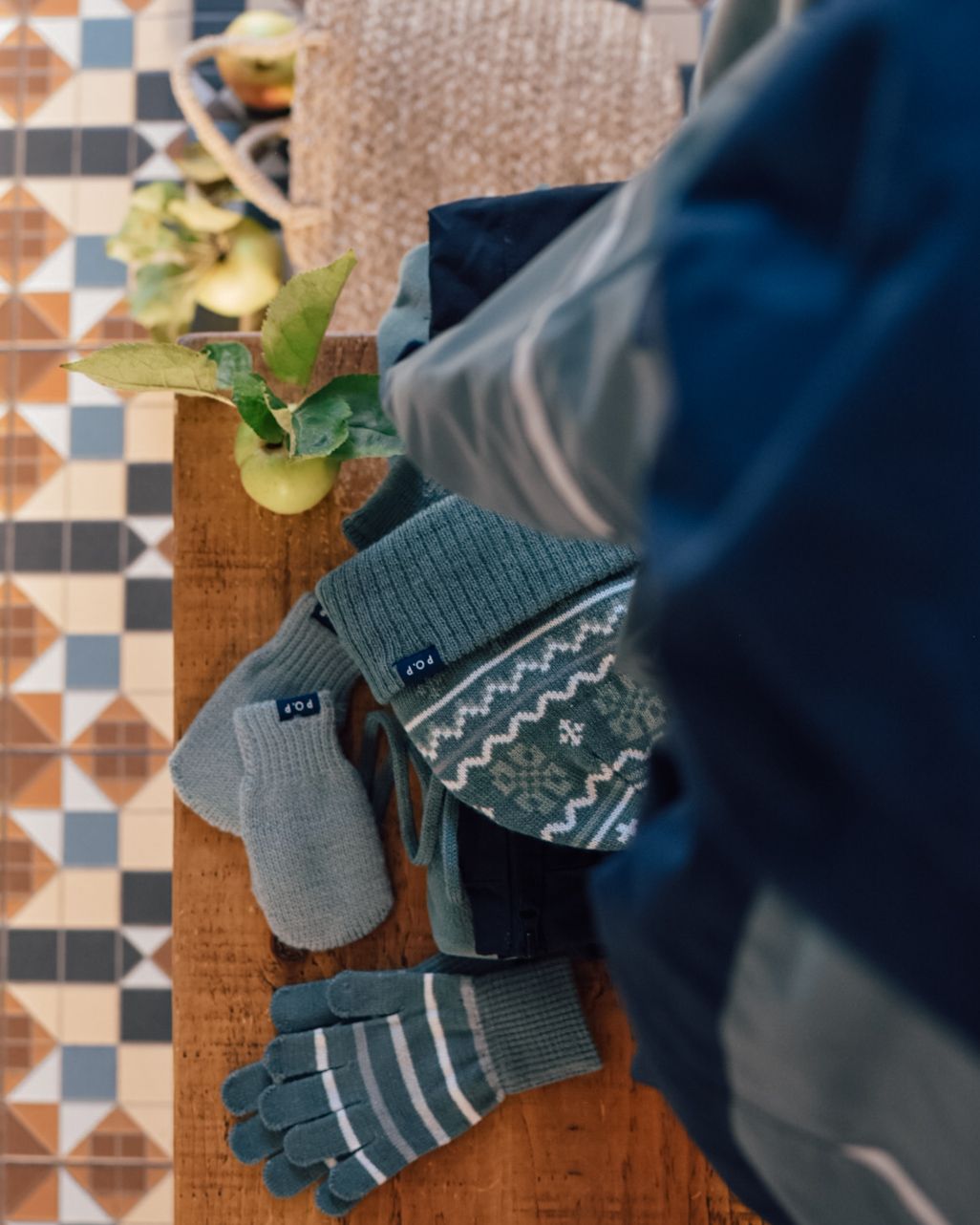 Wooly hat, gloves and mittens on a wooden top with coloured floor tiles in the background.