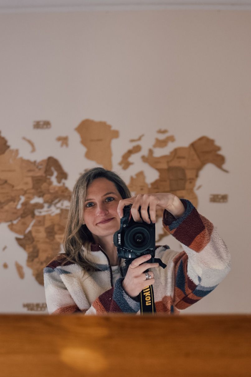 Laura holding a Nikon camera taking a self portrait in front of a mirror with a wooden map of the world on the wall in the background