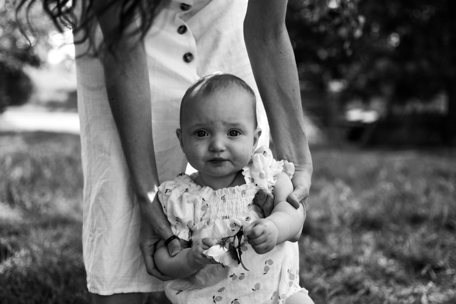 Black and white photo of a mothers arms supporting a baby as she sits in the garden