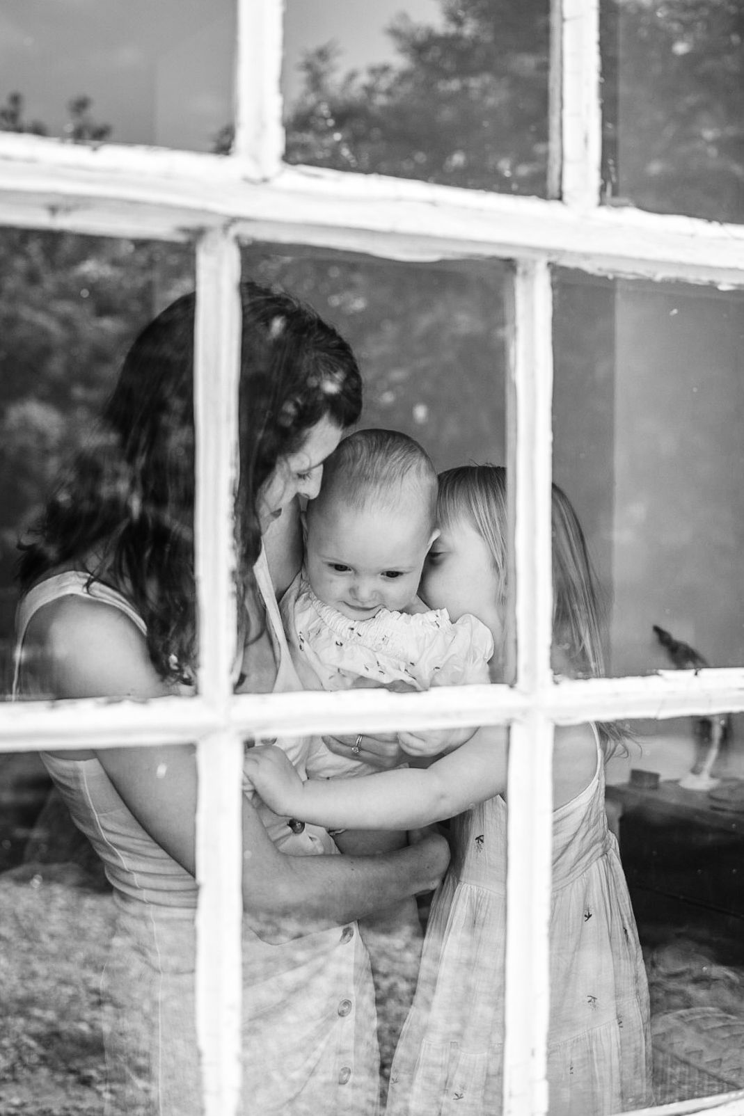 Mother and girl kissing baby in front of a window, seen from outside the window. Black and white