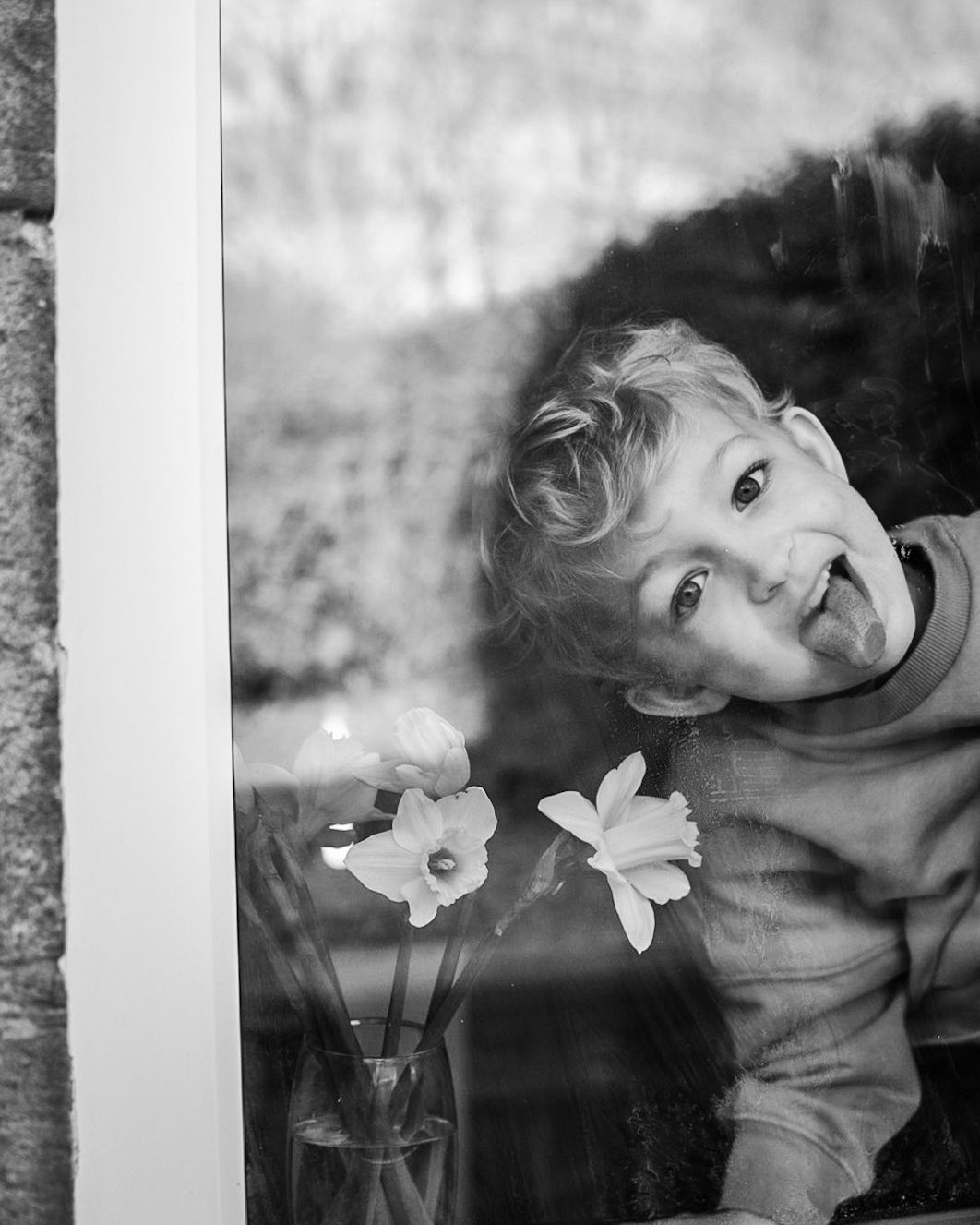 Black and white photo of boy looking through a window next to a vase with daffodils taken from outside the house.