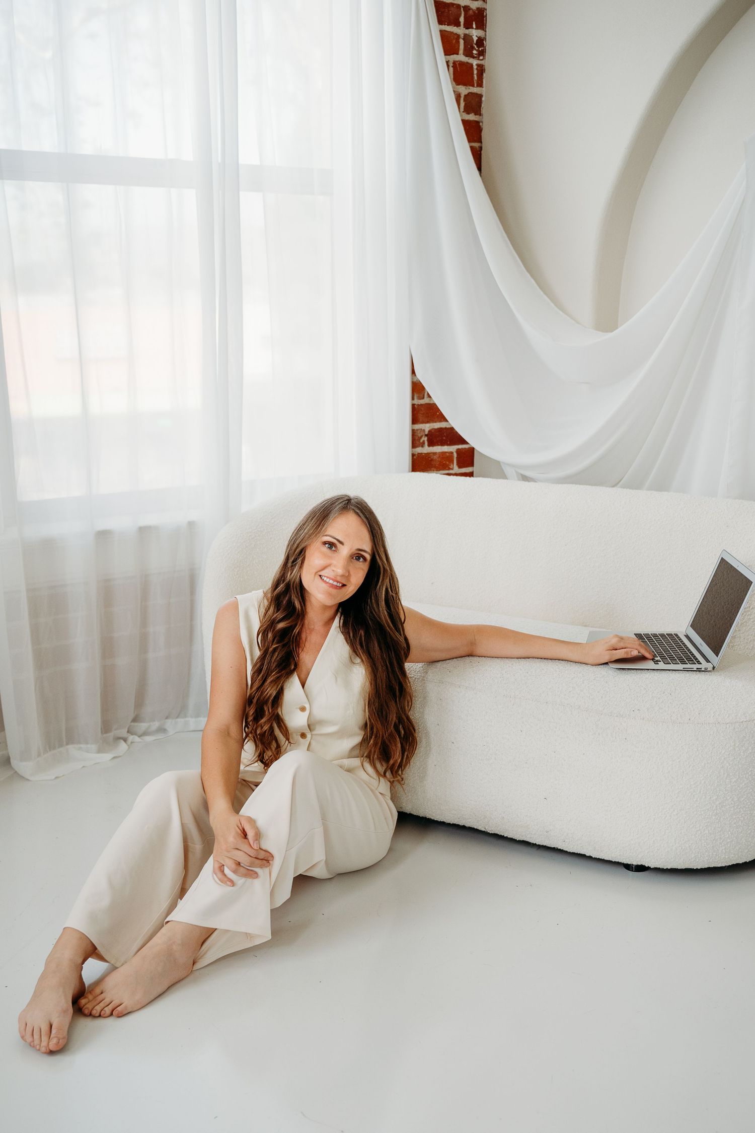 Happy barefoot woman with long wavy hair in cream outfit sitting on white floor beside sofa with laptop.