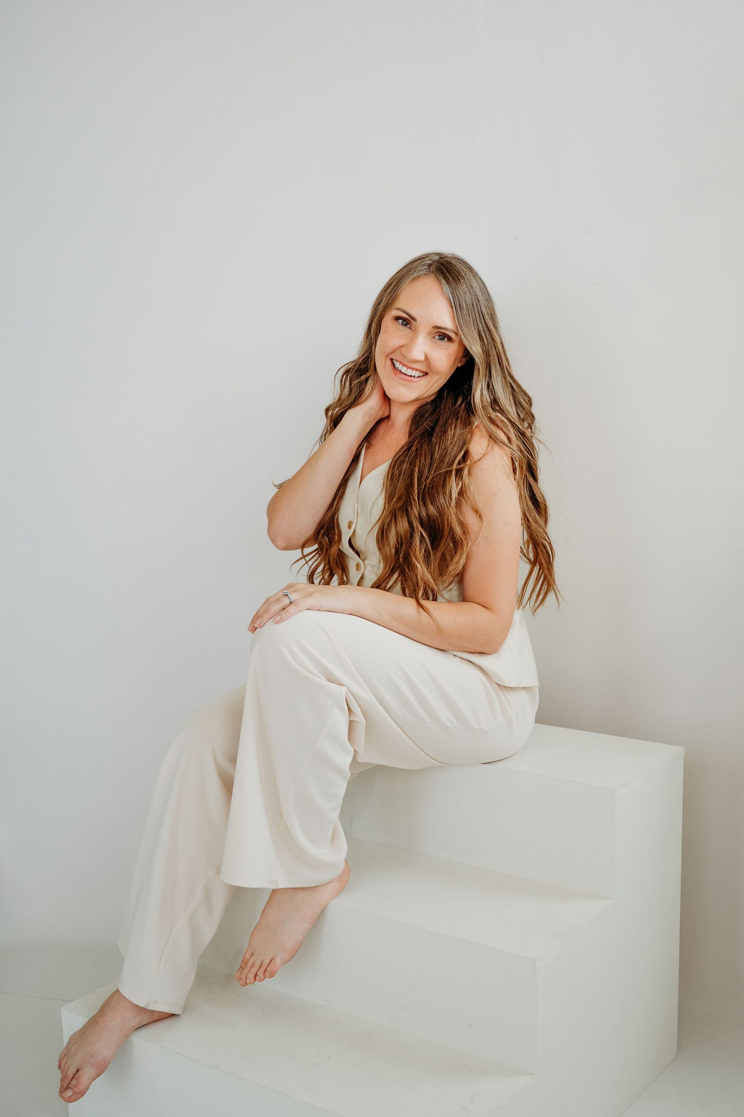 Cheerful barefoot woman with long wavy hair in cream outfit sitting on white steps in bright studio.