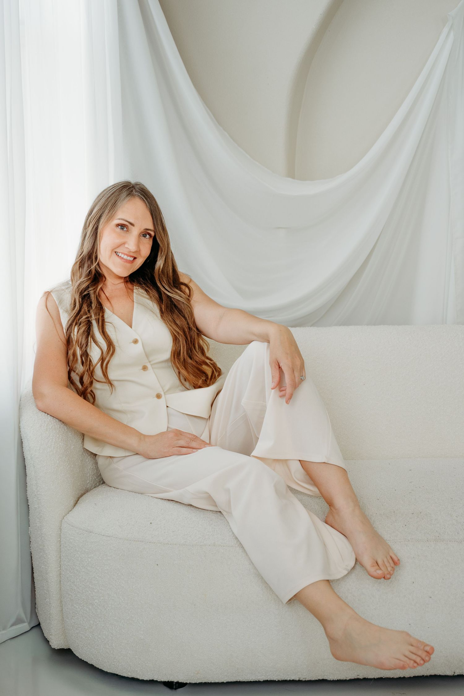 Smiling woman with long wavy hair sitting barefoot on white sofa wearing cream linen outfit in bright studio.