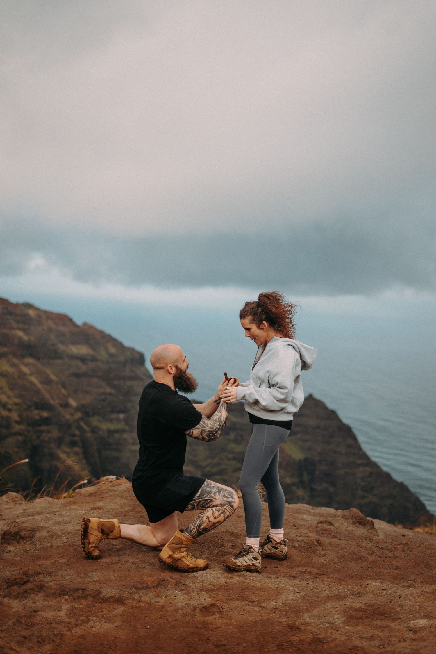 Kaua‘i engagement photoshoot on the Nā Pali Coast featuring a couple embracing at sunset with ocean views, tropical cliffs, and  beach