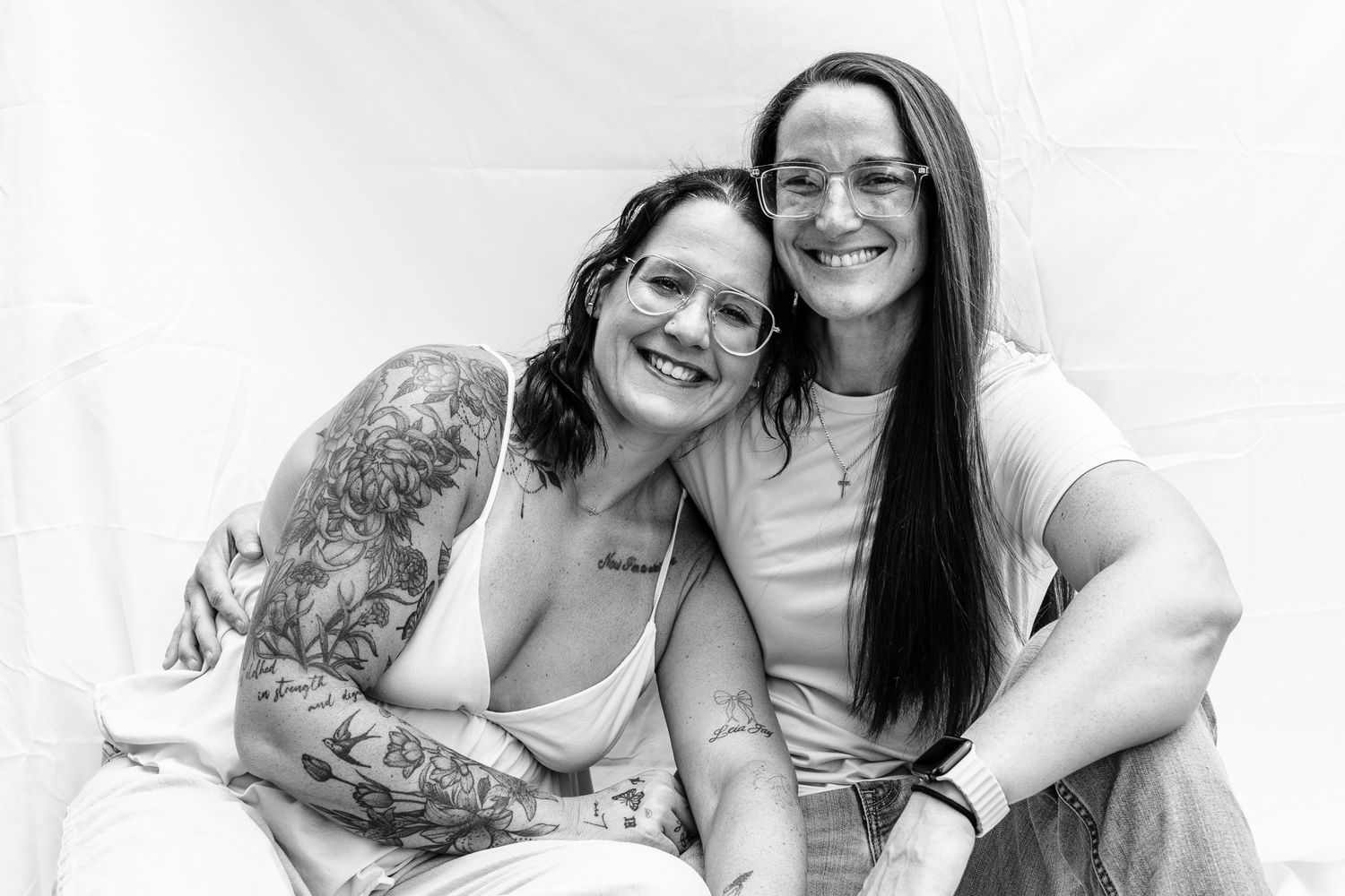 Two smiling women pose together in a black and white photo, one with tattoos, both looking happy.
