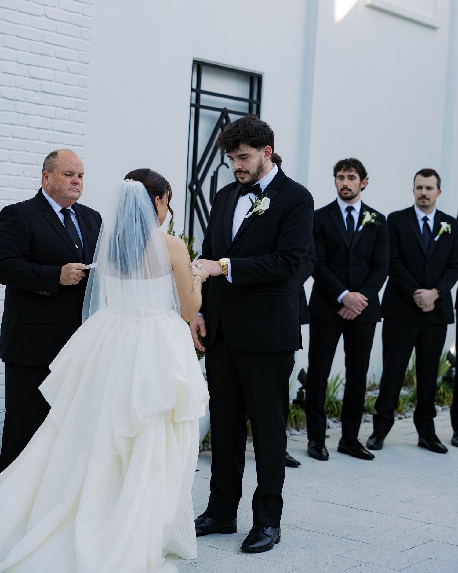 Wedding party in formal attire poses against white brick wall during ceremony.