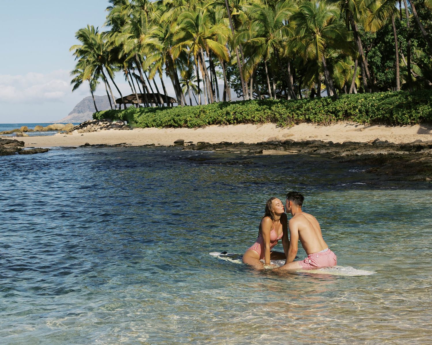 Palm trees line a pristine beach cove with two people floating on surfboards in turquoise waters.