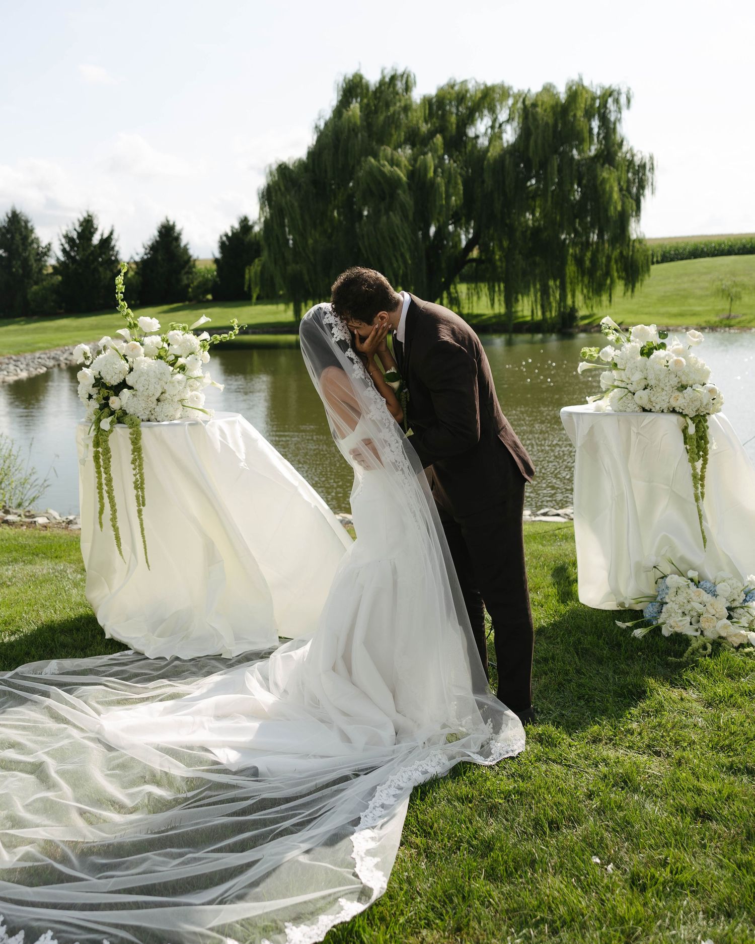 Bride and groom share a romantic kiss at outdoor wedding ceremony by a pond, surrounded by elegant white floral arrangements.