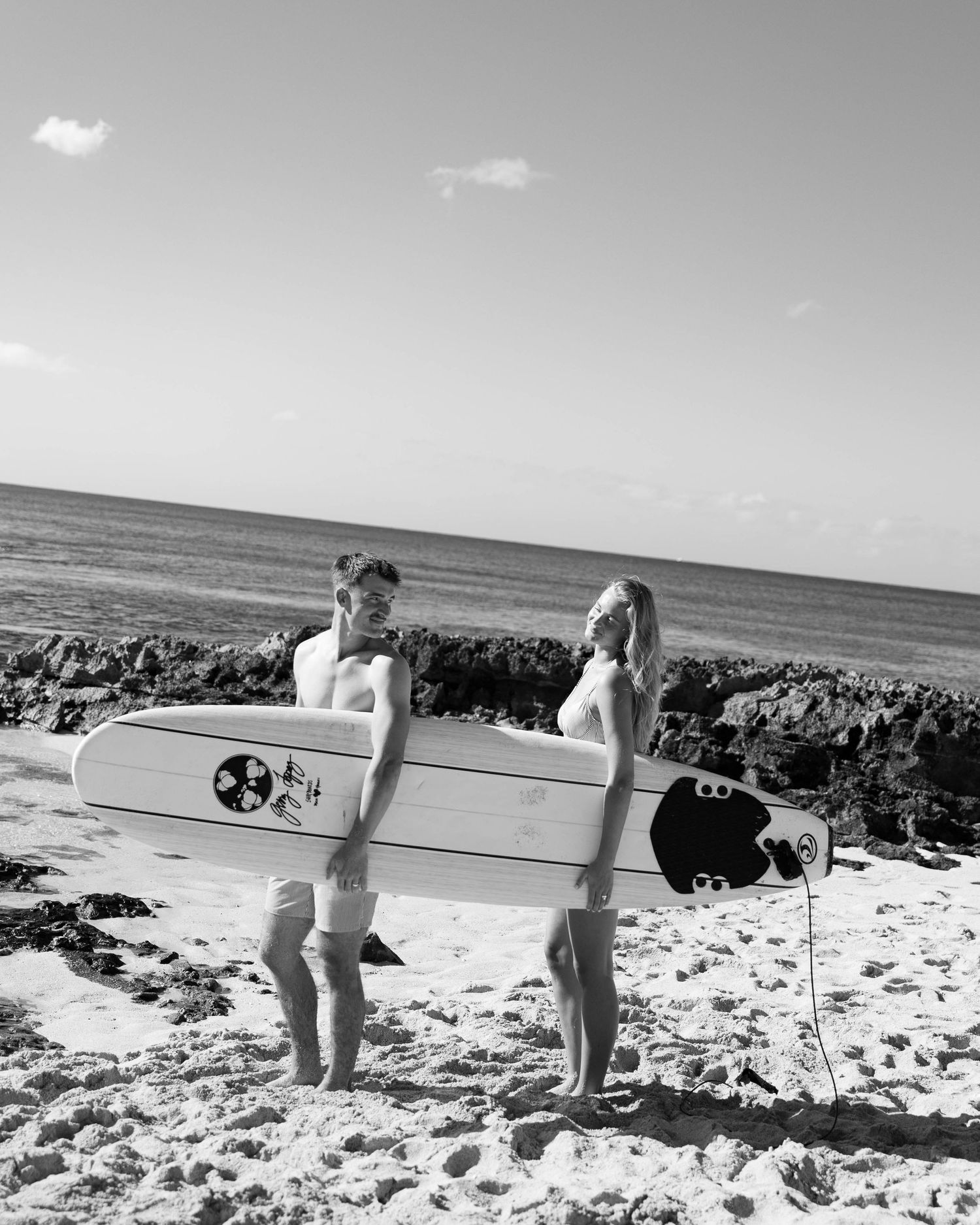 Two surfers carrying a longboard across a rocky beach in black and white.