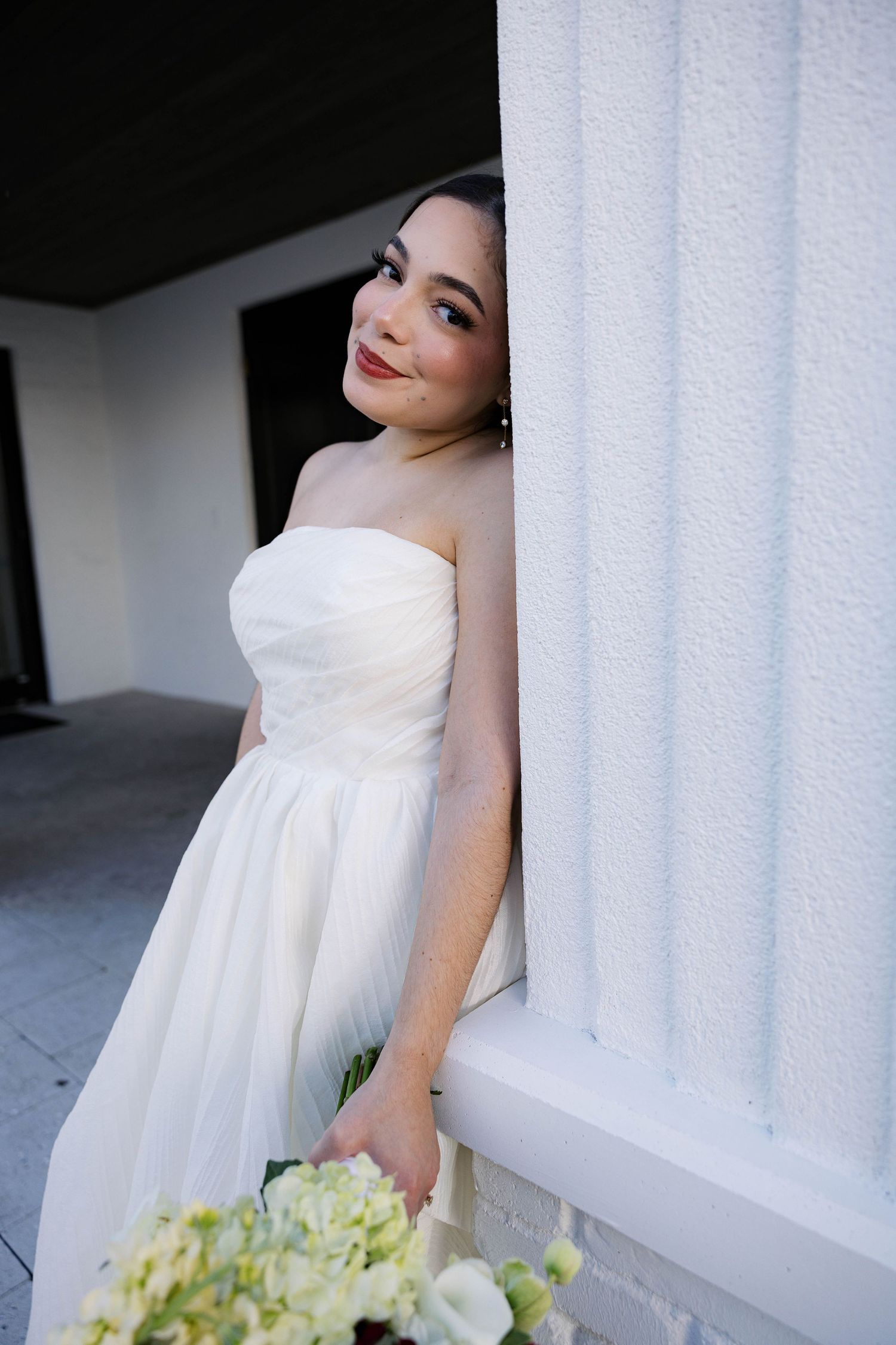 A bride in a flowing white dress poses elegantly against white columns with soft lighting and greenery.