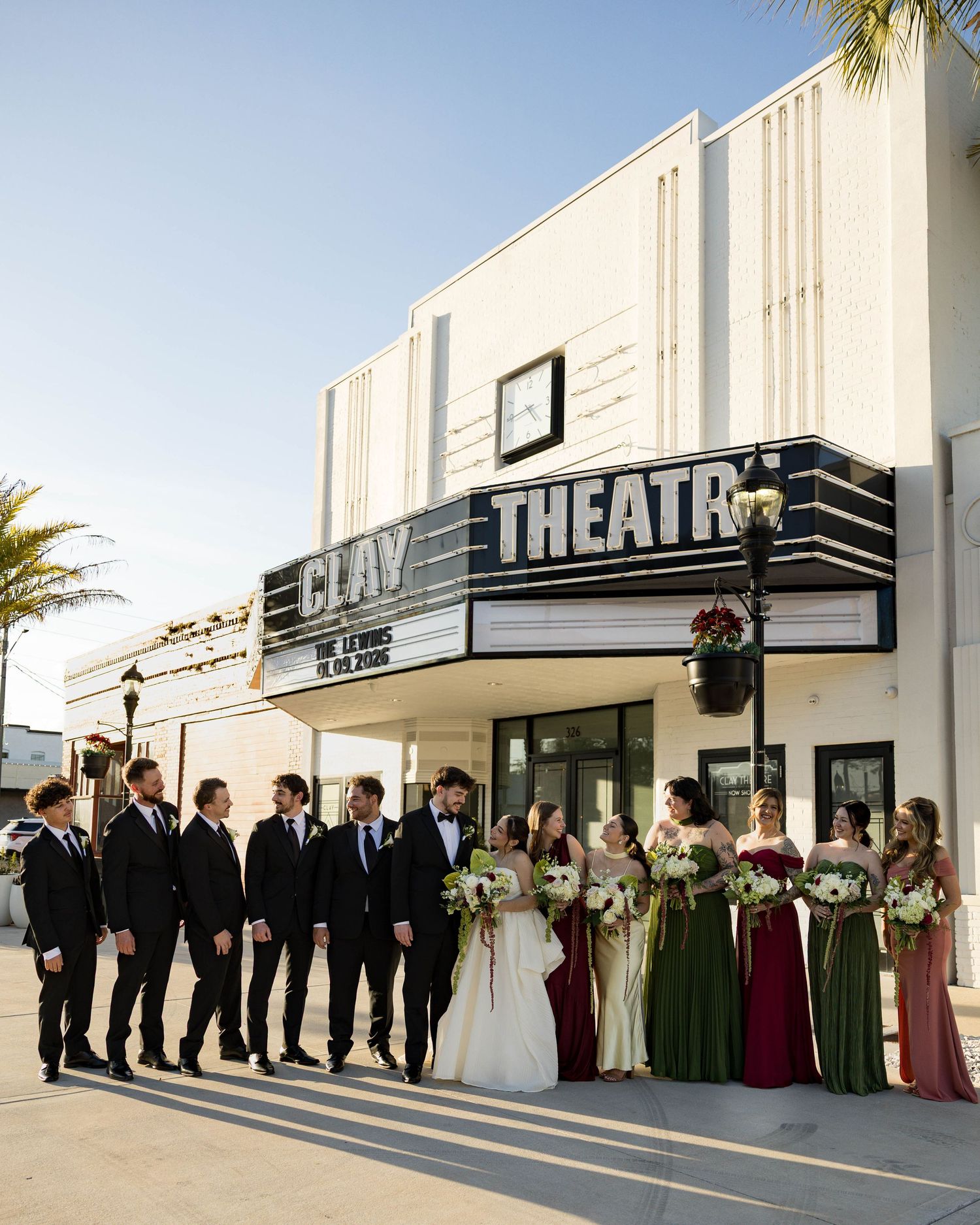 Wedding group portrait outside historic theatre with bridesmaids in jewel-toned dresses.
