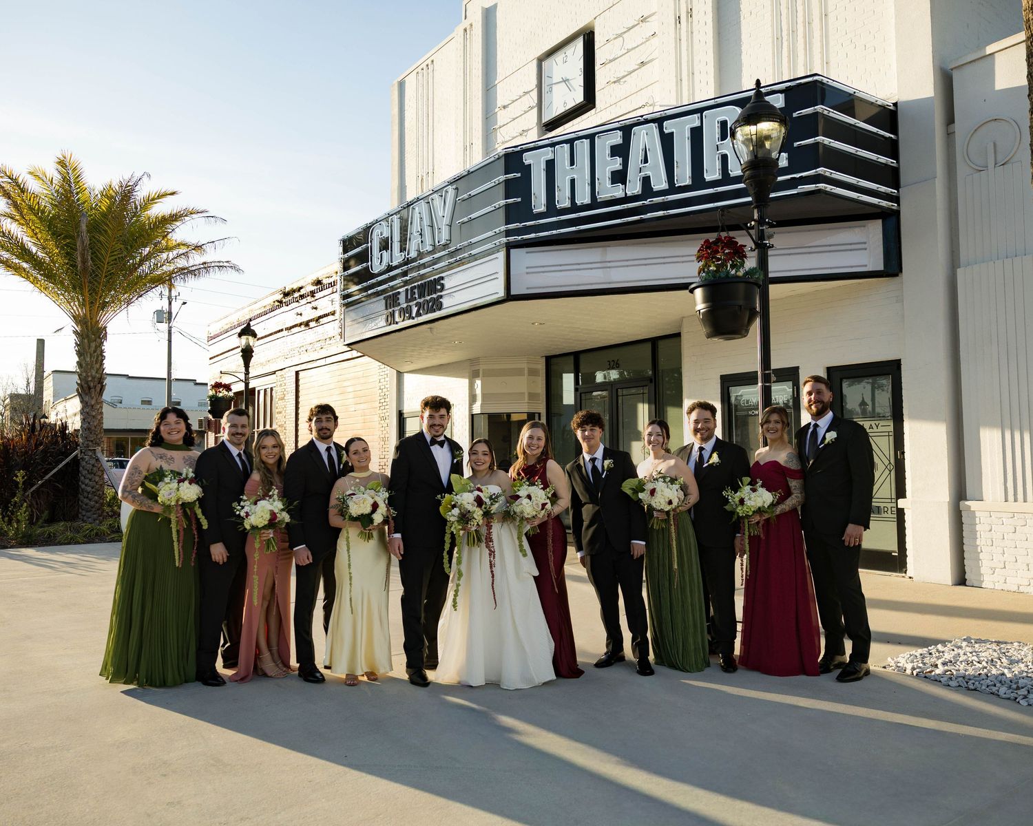 Wedding party poses in front of a vintage theater with bridesmaids in green and burgundy dresses holding white bouquets.