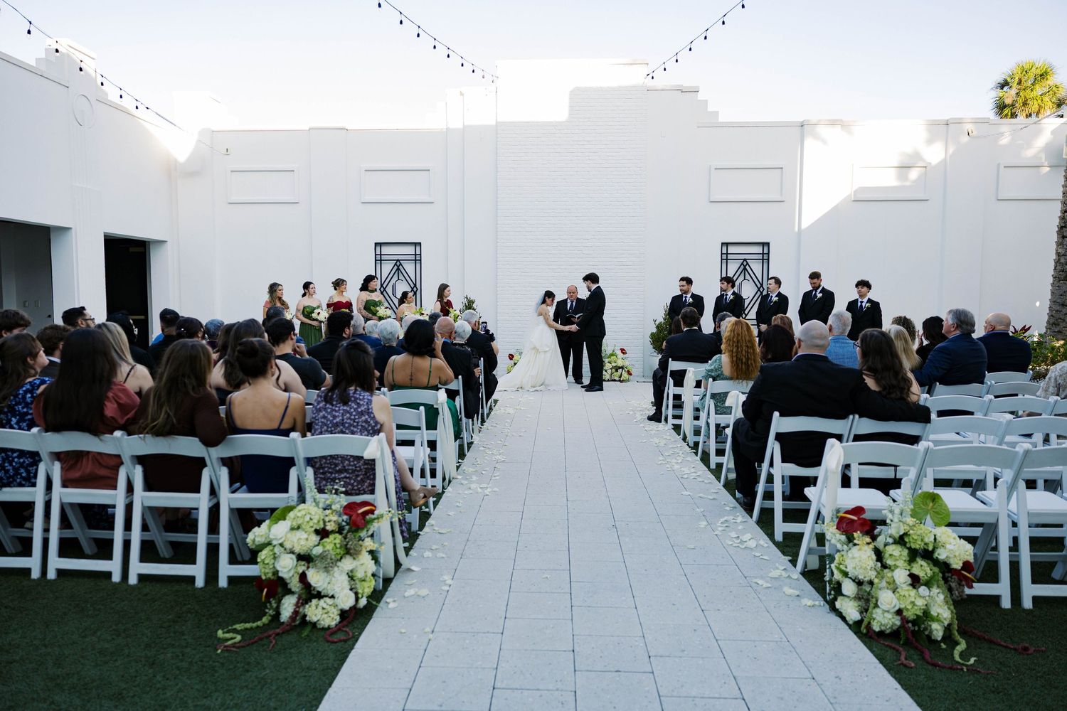 Outdoor wedding ceremony setup with white chairs and floral arrangements along the aisle.