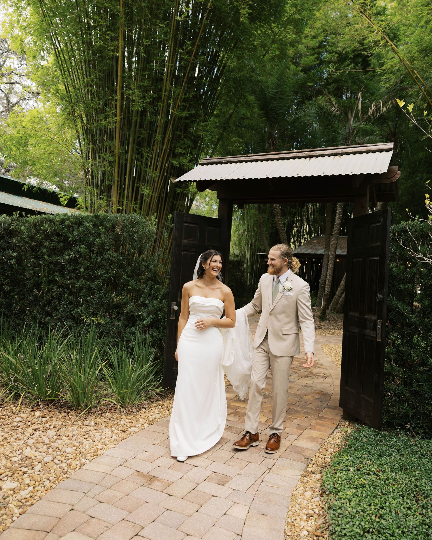 Couple walks along bamboo-lined garden path in wedding attire.